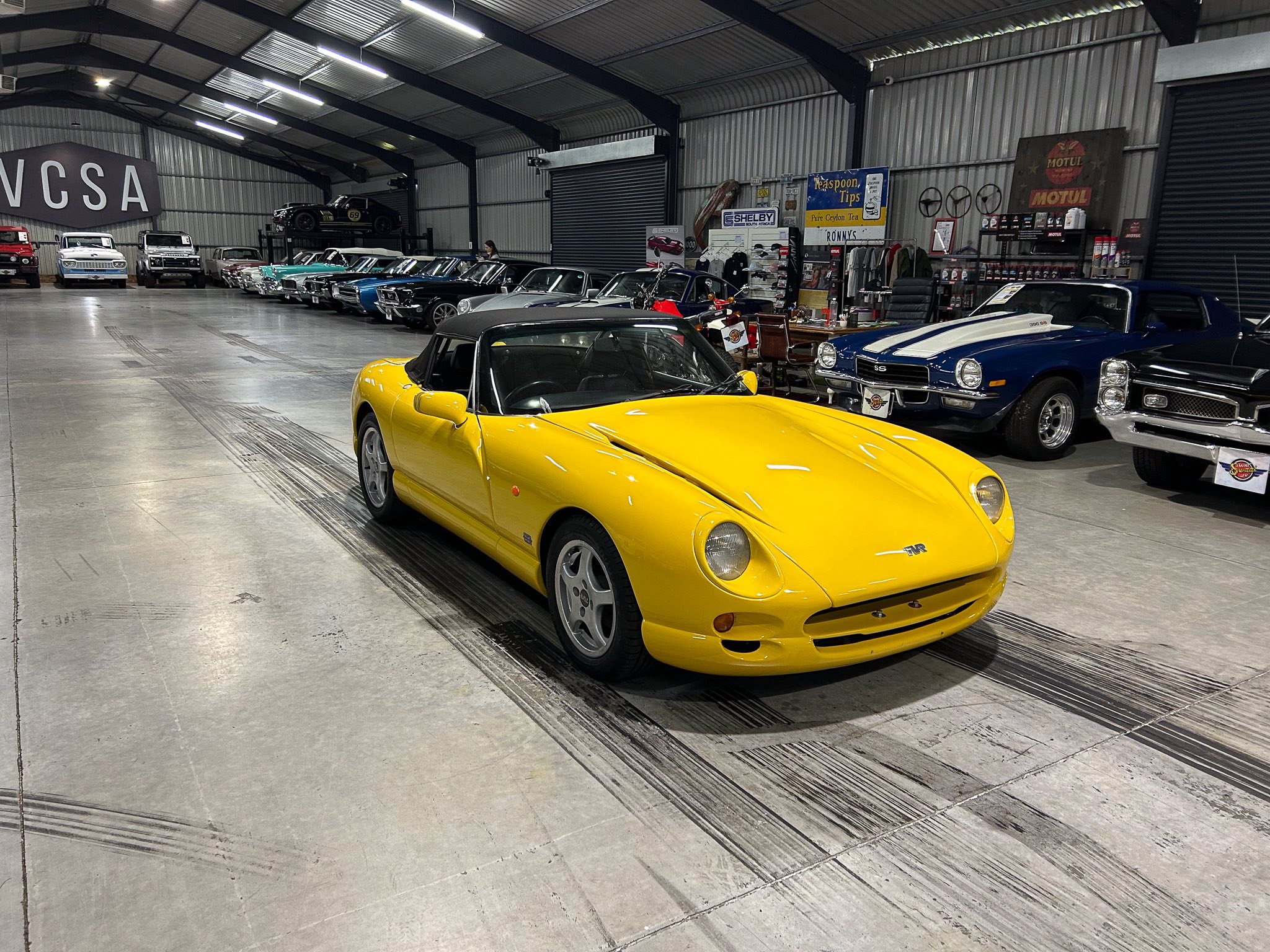 Indoor car showroom with a bright yellow convertible in the foreground and a row of classic cars along the wall inside a large warehouse-like space