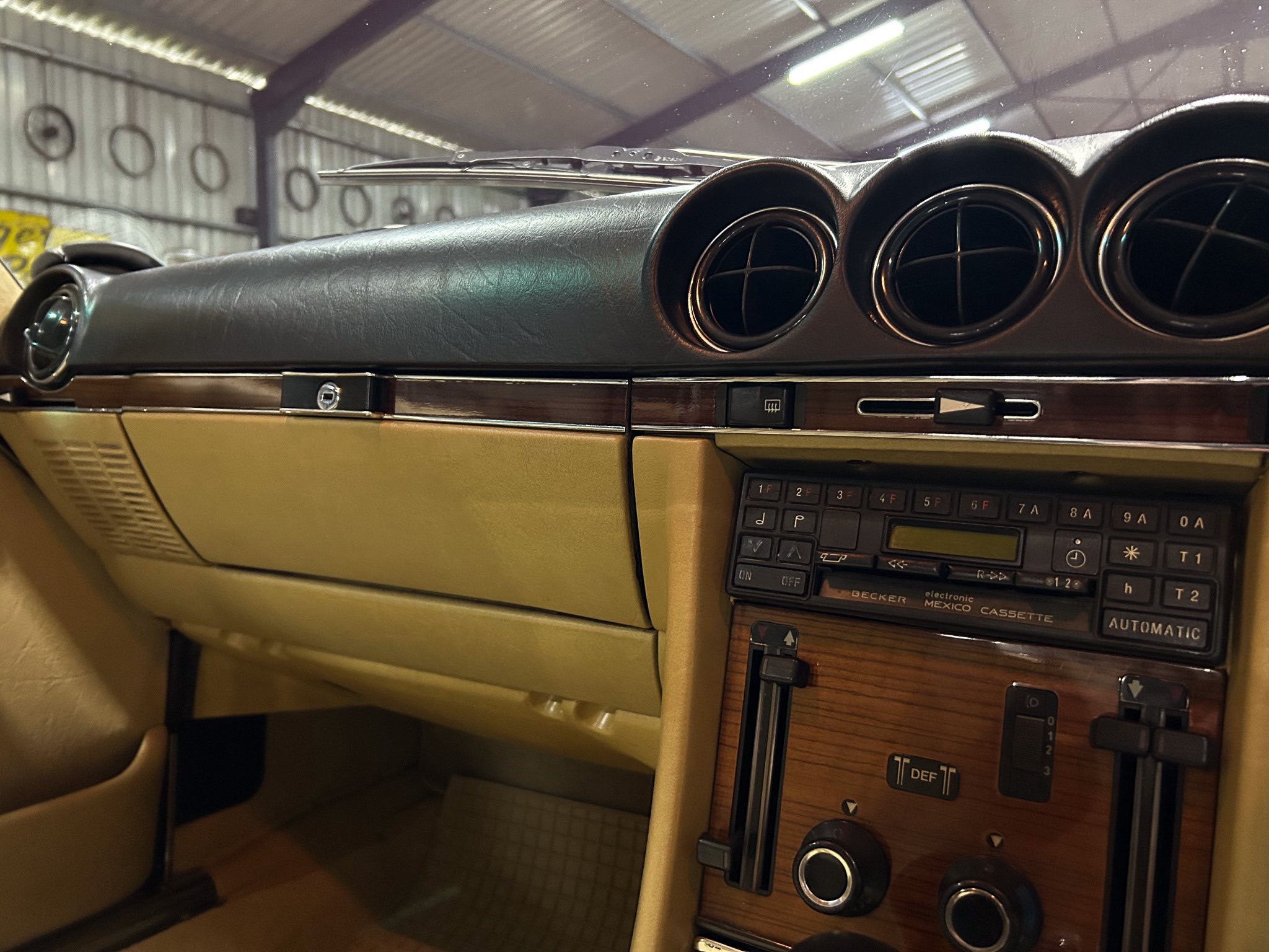 Beige car interior featuring a wood-grain center console with three round air vents and vintage climate/radio controls.