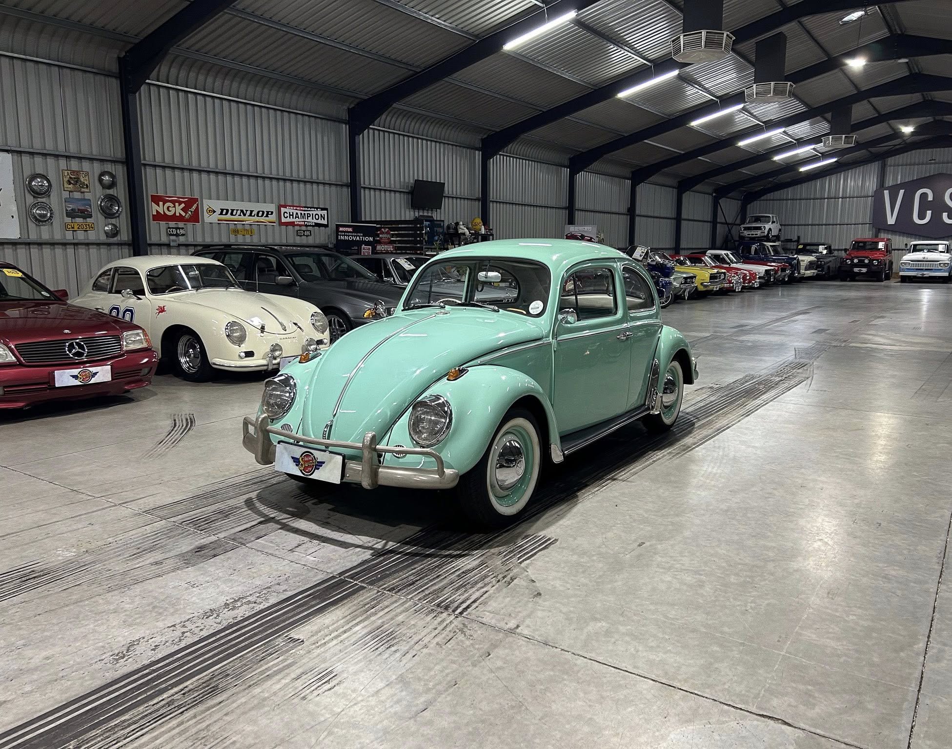 Indoor classic car showroom with mint green vintage Volkswagen Beetle in foreground and rows of classic cars along the wall; banners and workshop gear visible in background