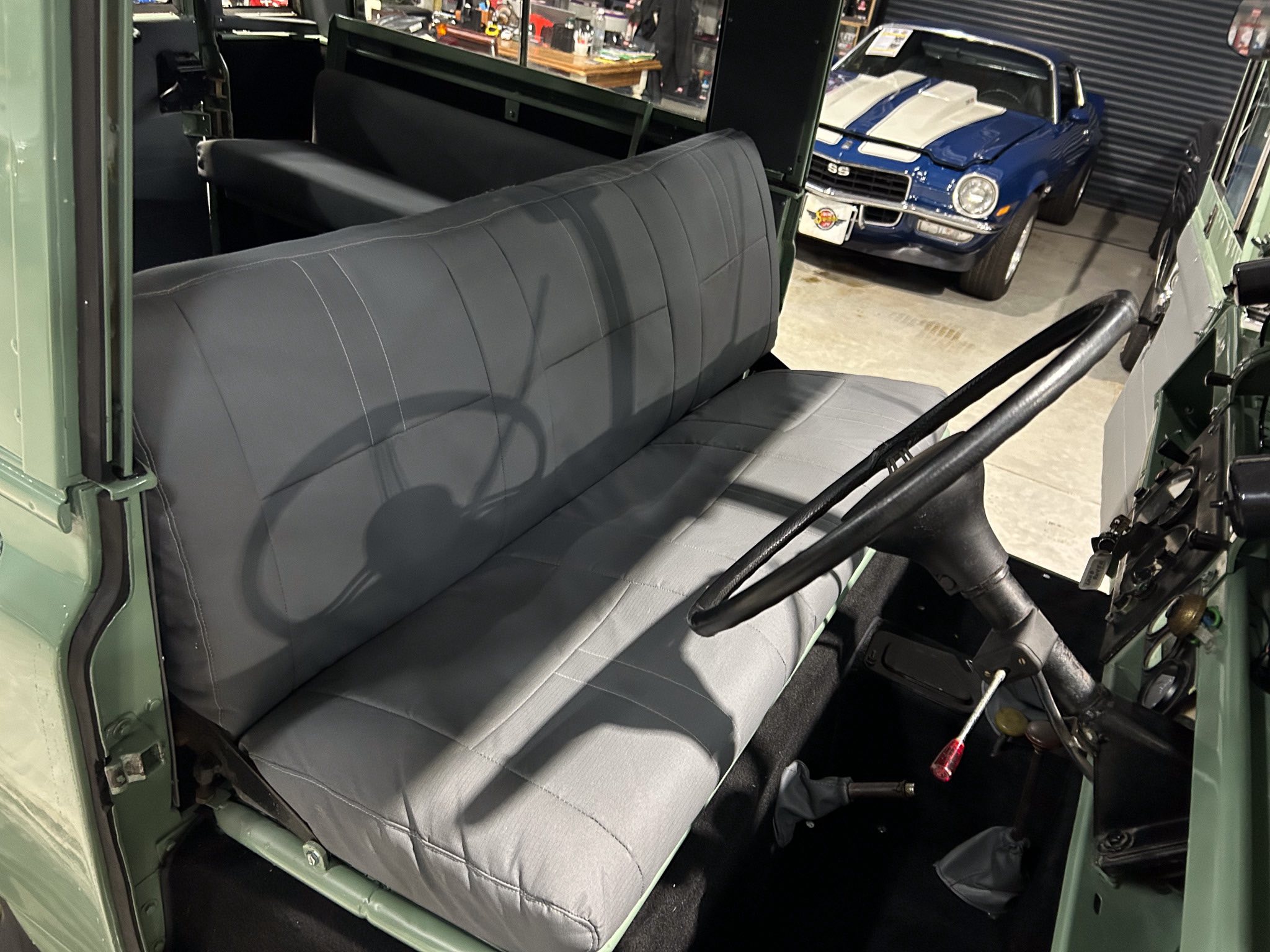 Interior of a vintage green vehicle with a gray vinyl bench seat and steering wheel visible from the driver's side door