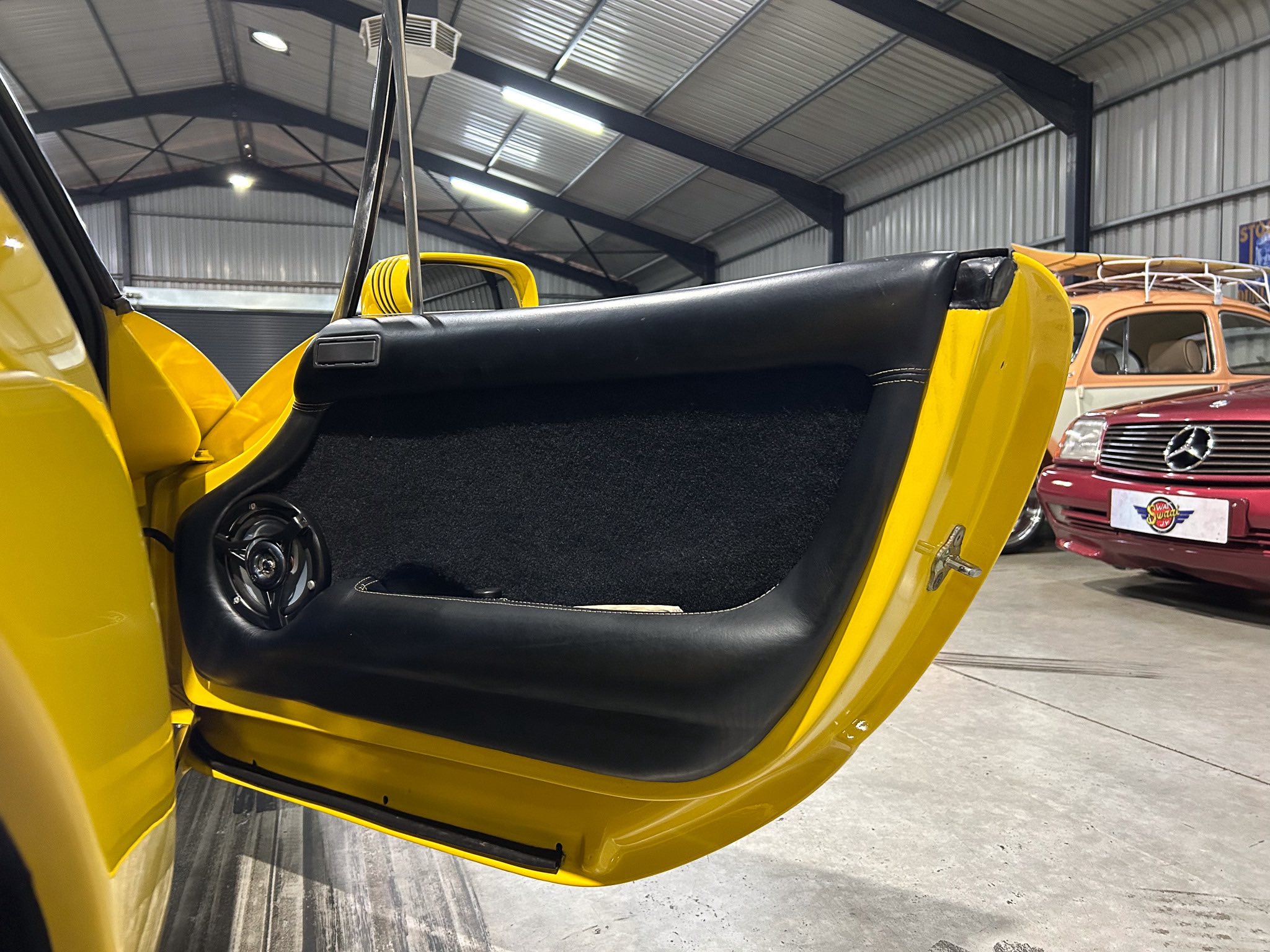 Close-up of a yellow sports car with its door open in a garage, showing black leather interior and a gull-wing-style door; other vintage cars visible in the background.