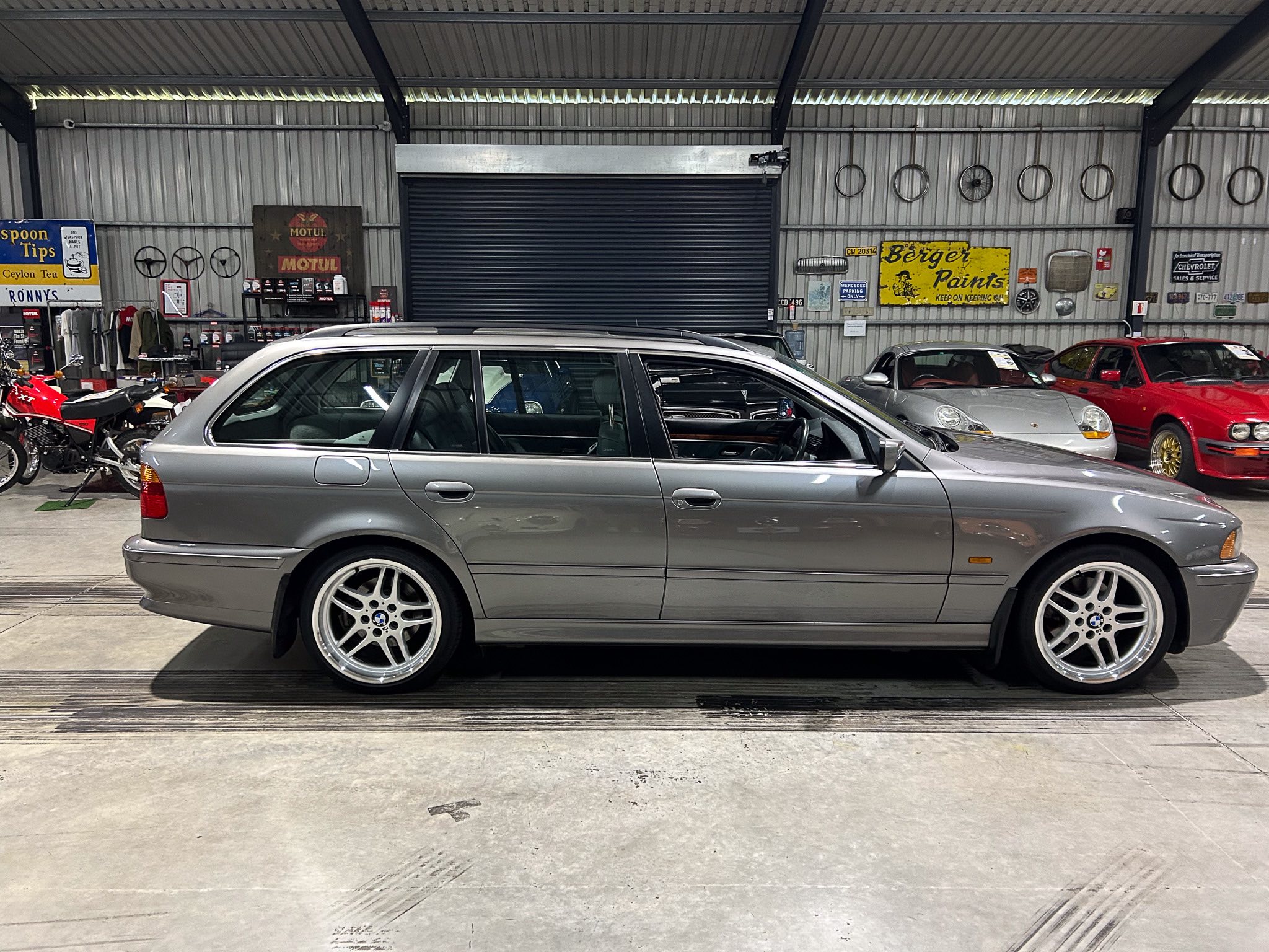 Silver BMW wagon in a showroom/garage with other cars and vintage signs on corrugated metal walls in the background.
