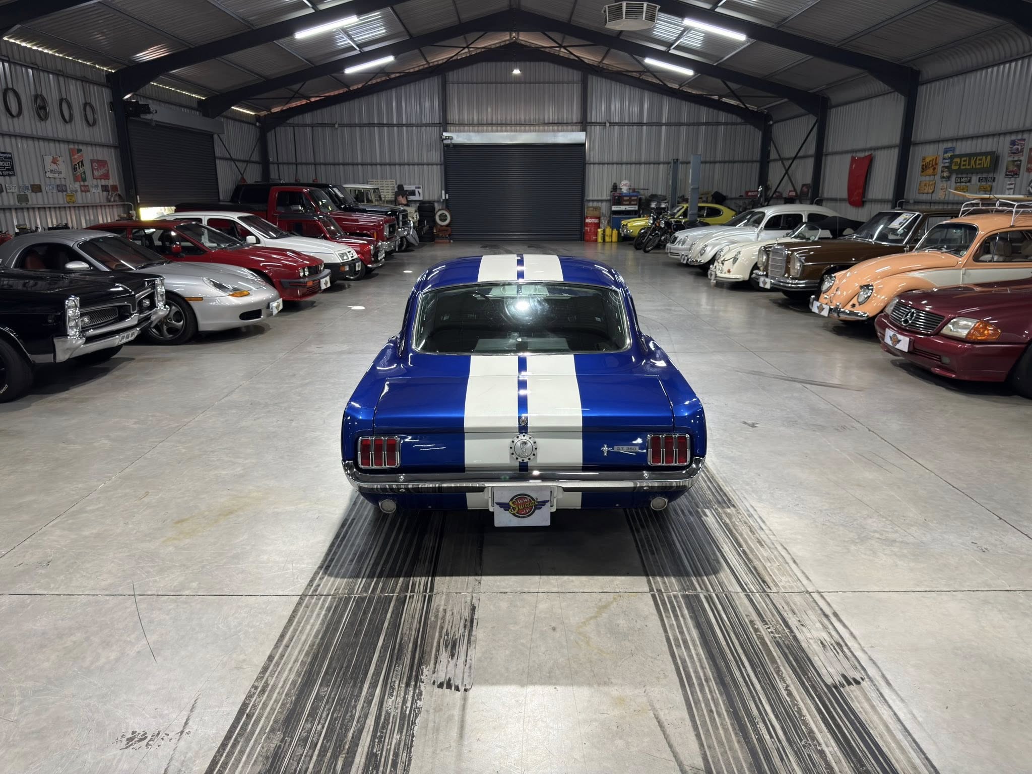 Blue vintage car with white race stripes is centered in a large metal garage showroom filled with rows of classic cars on both sides.