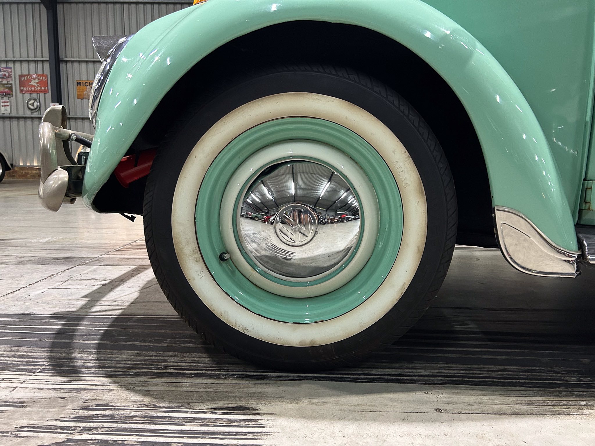 Close-up of a vintage car wheel with a mint-green fender, whitewall tire, and chrome hubcap reflecting the surroundings in a showroom/Garage.