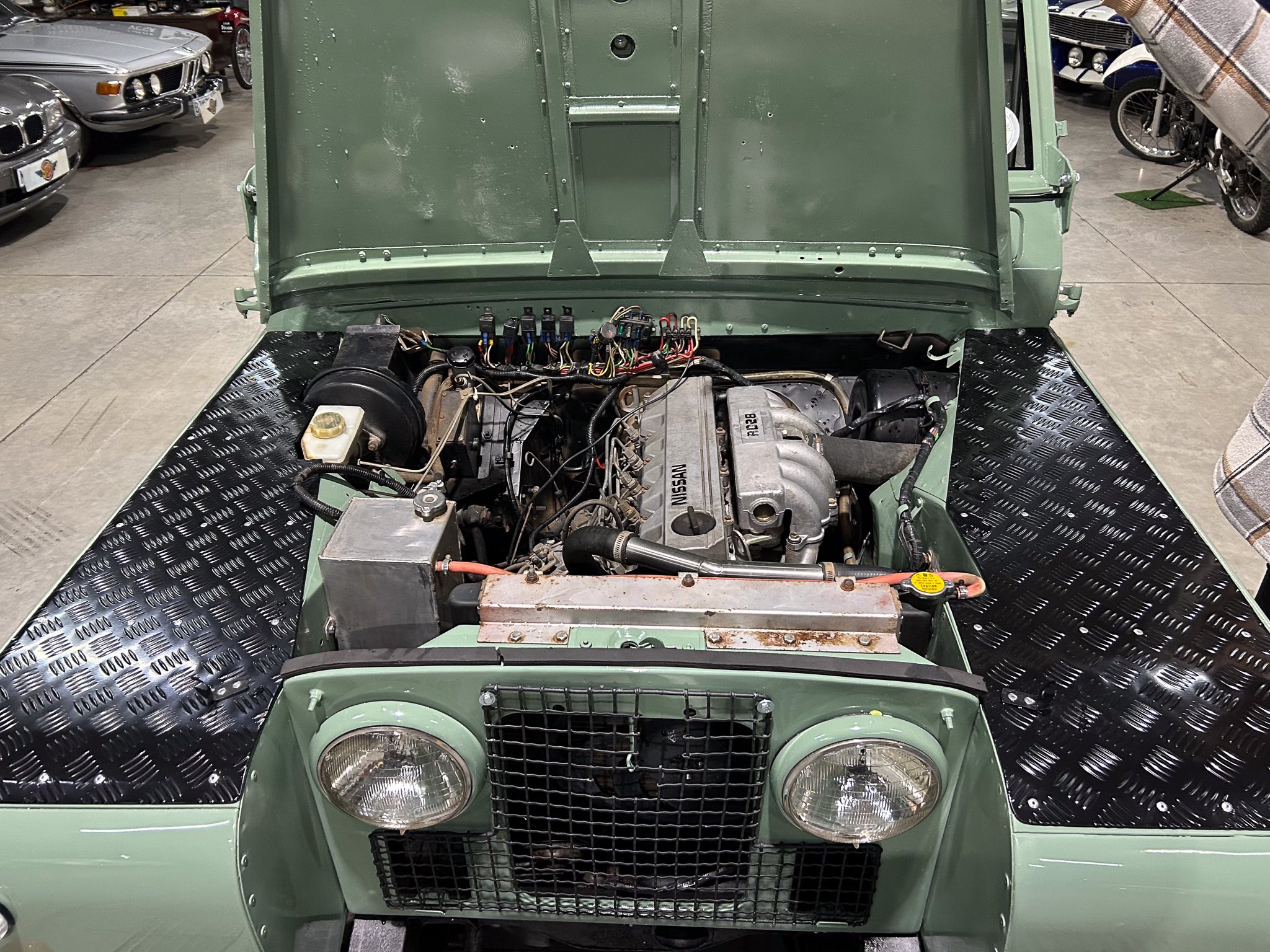 Front view of a green vintage Nissan with hood open, engine bay exposed and wiring visible on a diamond-plate display floor. Cars in background show an indoor showroom.