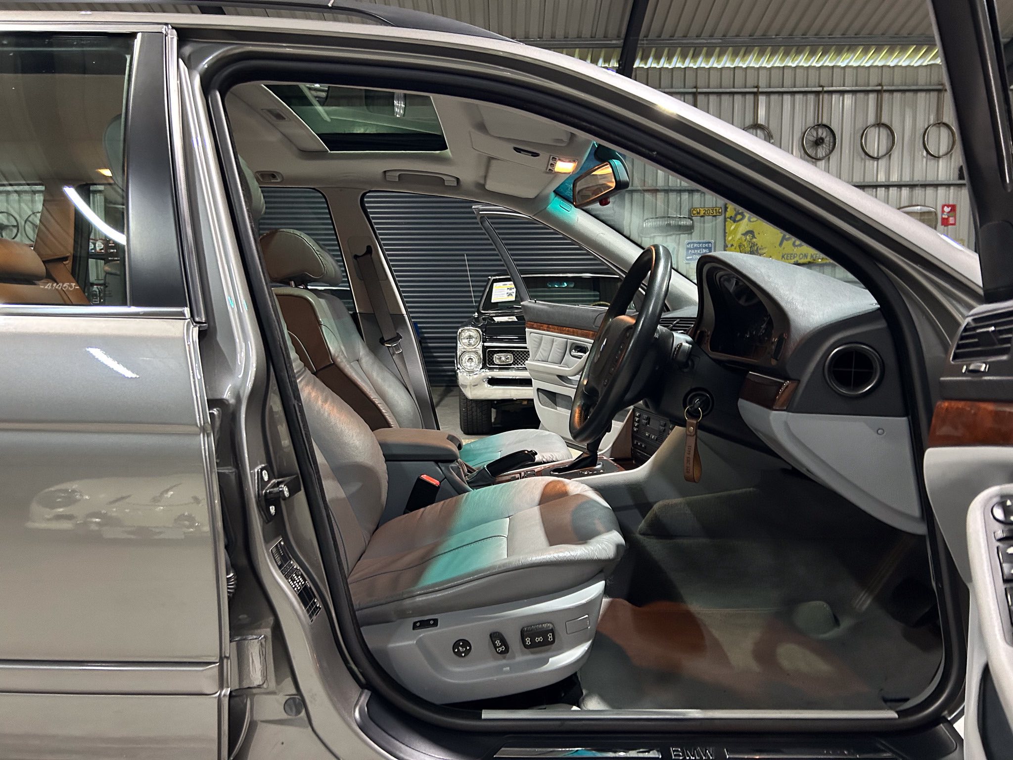 Open SUV door to reveal tan leather interior with wood trim, steering wheel, dashboard, sunroof, and a second car in a garage backdrop.