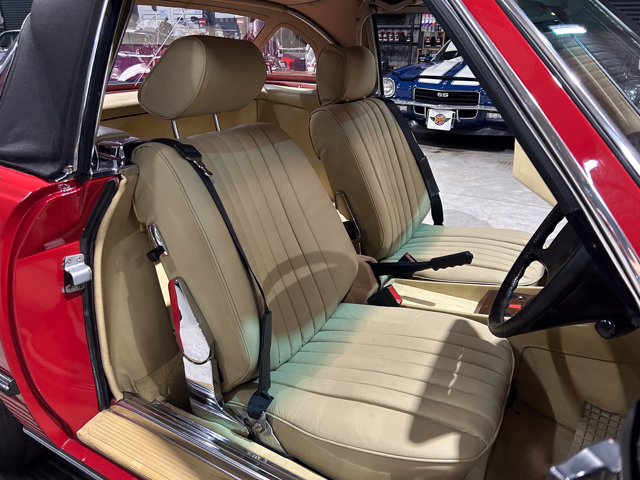 Inside a red car showing tan leather seats (front and rear) with seat belts, door open, showroom visible outside.