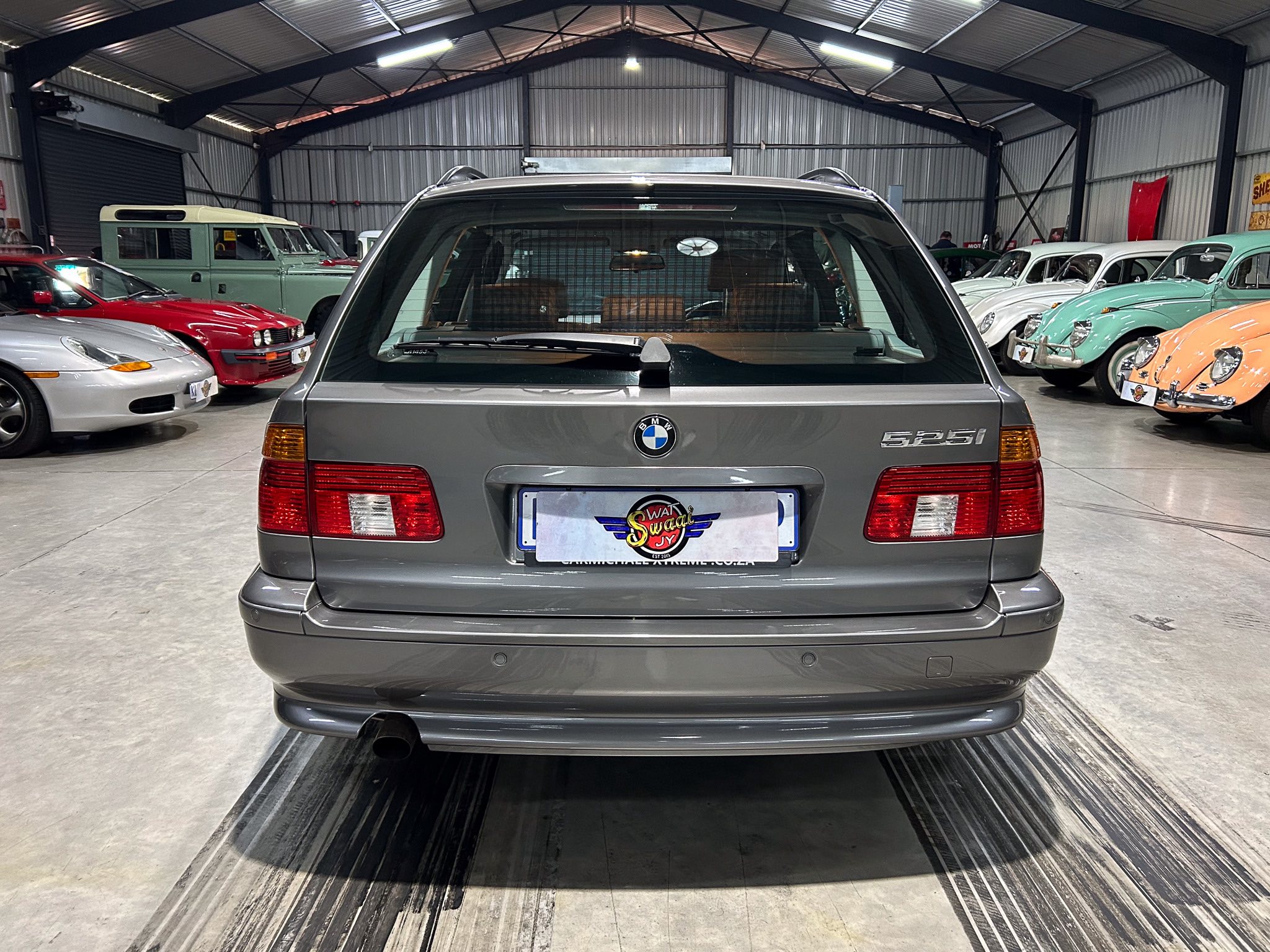 Rear view of a gray BMW 525i in a showroom with vintage cars on both sides of the space.