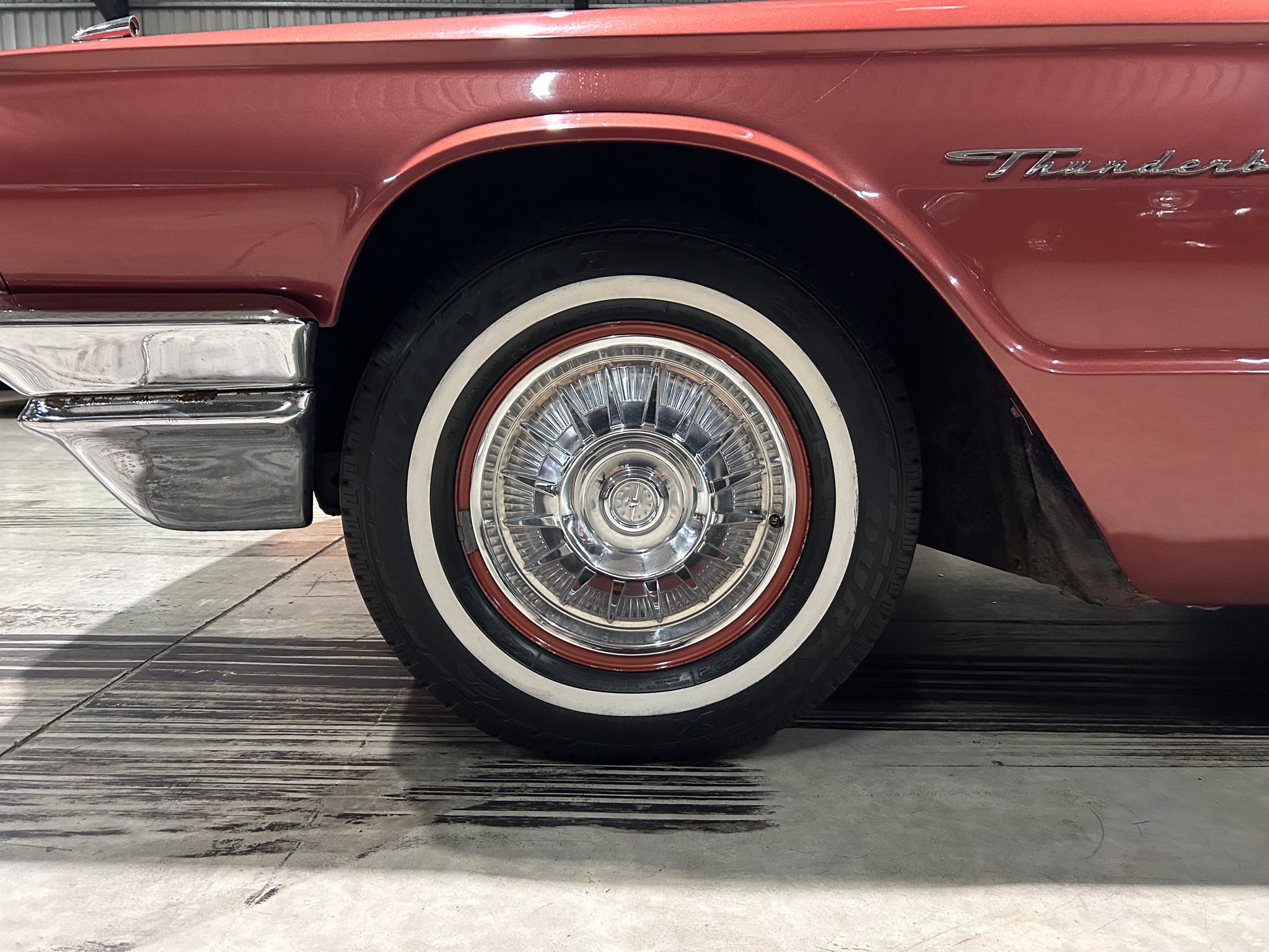 Close-up of a red vintage car's front fender showing a wheel, whitewall tire, and Thunderbird script badge.