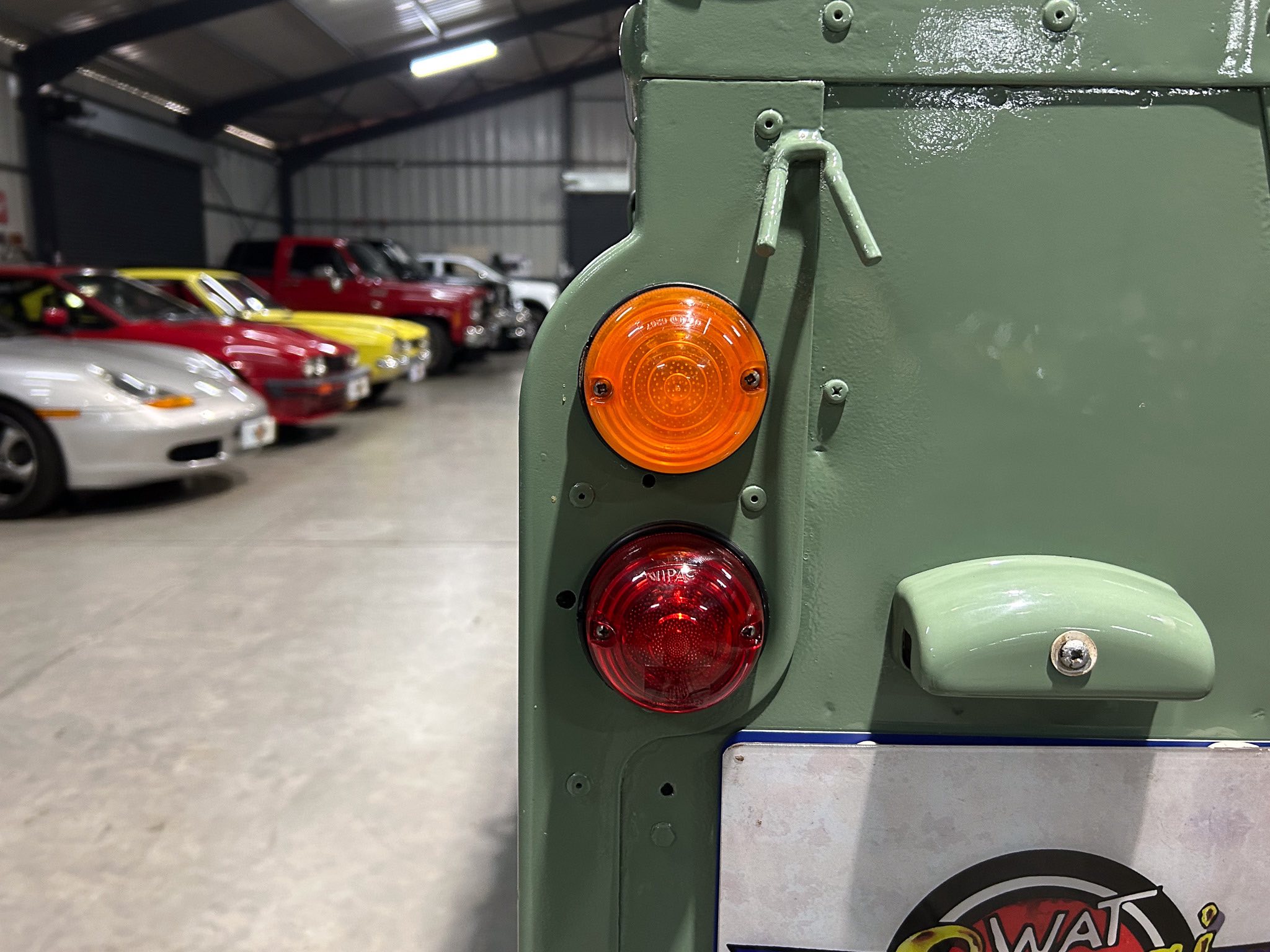 Close-up of a green vehicle's taillights with a row of colorful cars in a workshop background.