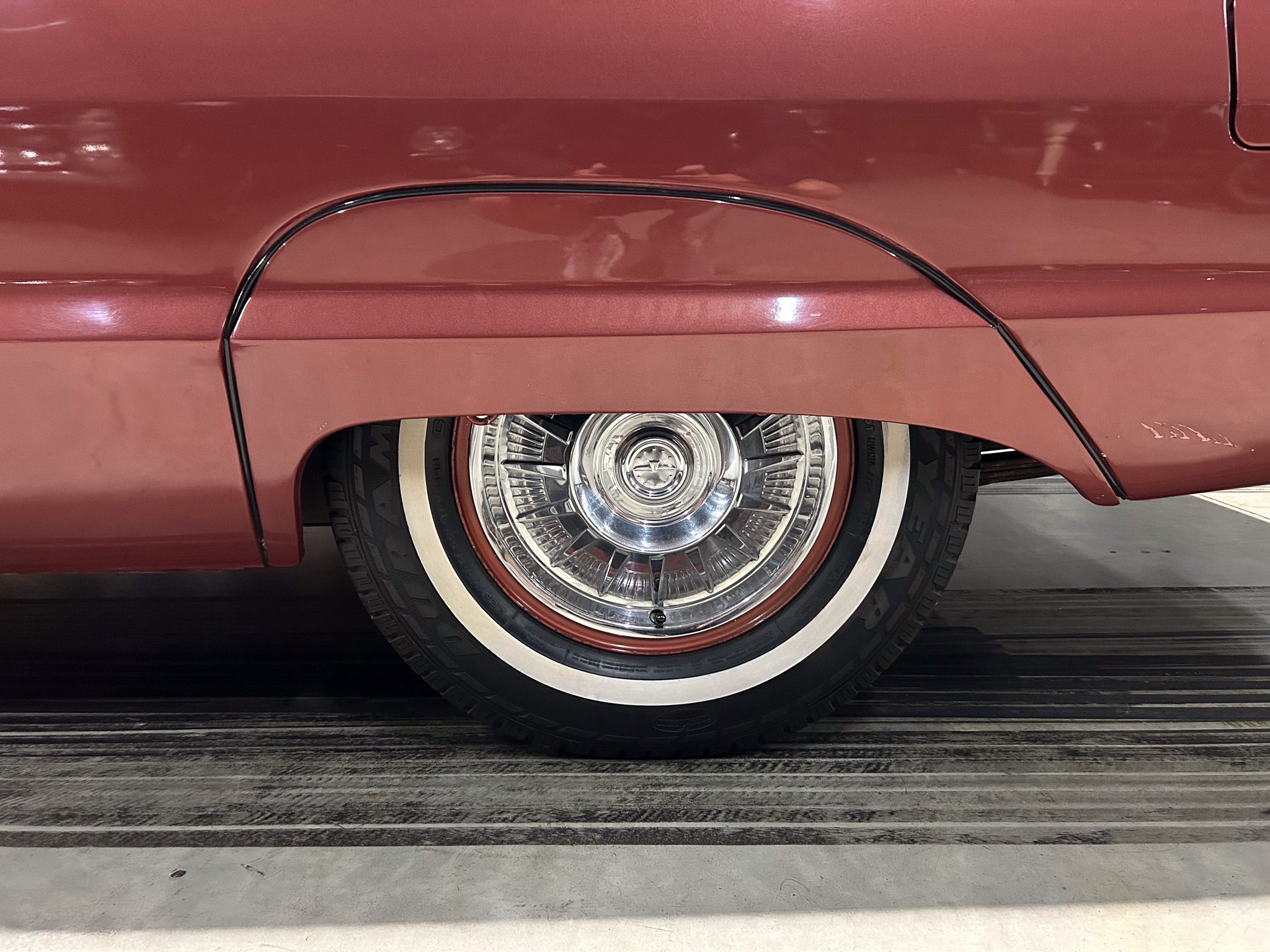 Close-up of a vintage car wheel with chrome hubcap and whitewall tire, set in a red fender, reflecting light.**NOTE**: Keep concise and informative for screen readers.
