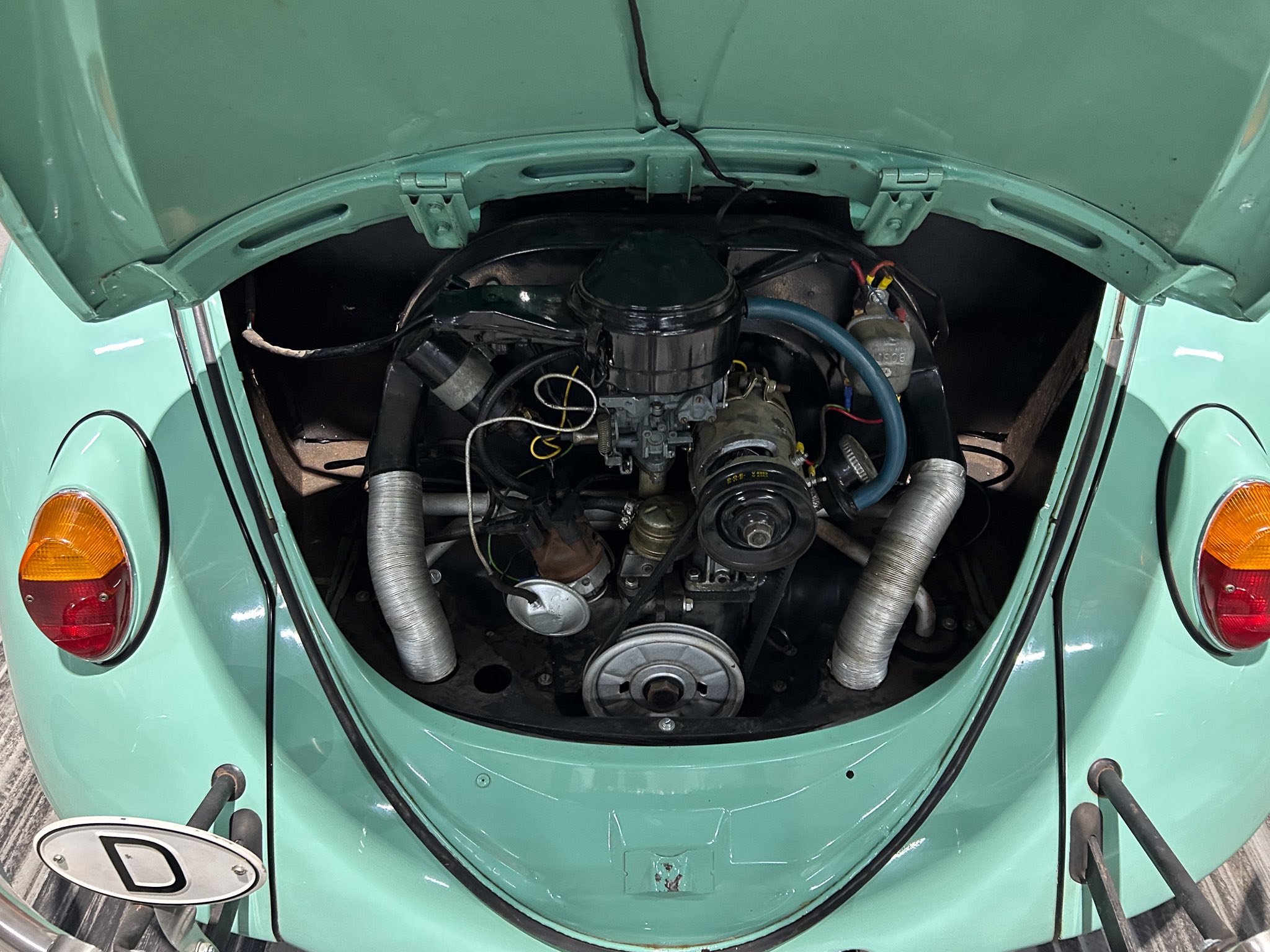 Rear engine bay of a teal vintage car with exposed hoses, wires and carburetor components visible under the open hood