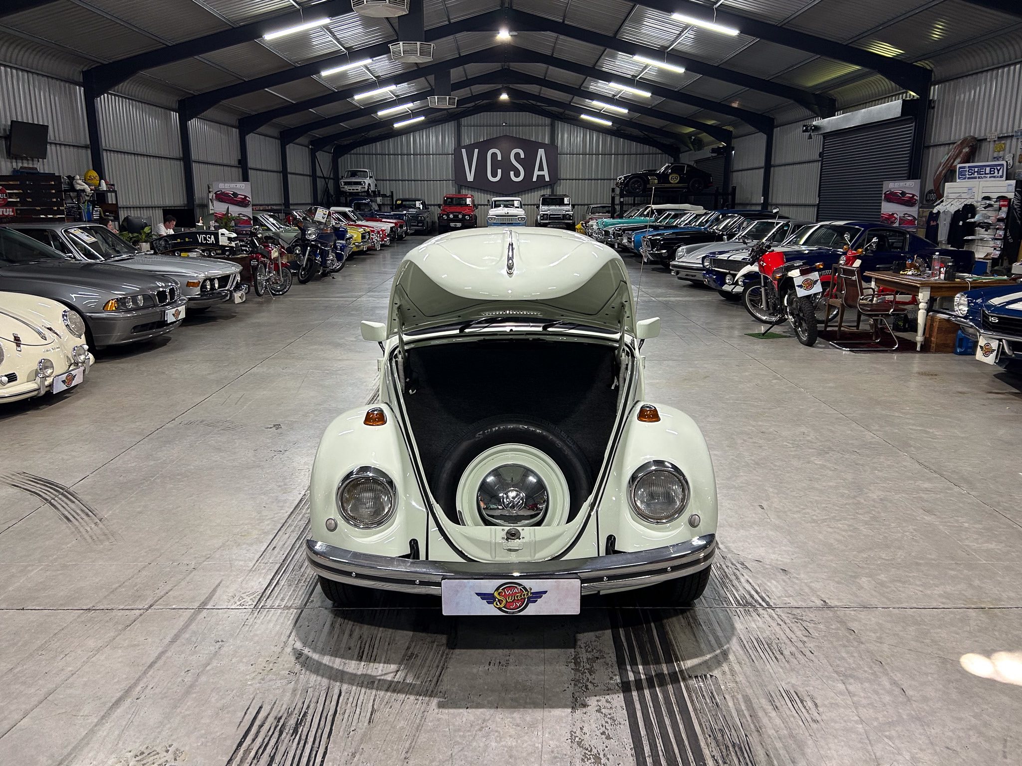 Front view of a pale green vintage car with its hood open inside a showroom filled with rows of classic cars and a large VCSA sign in the background.