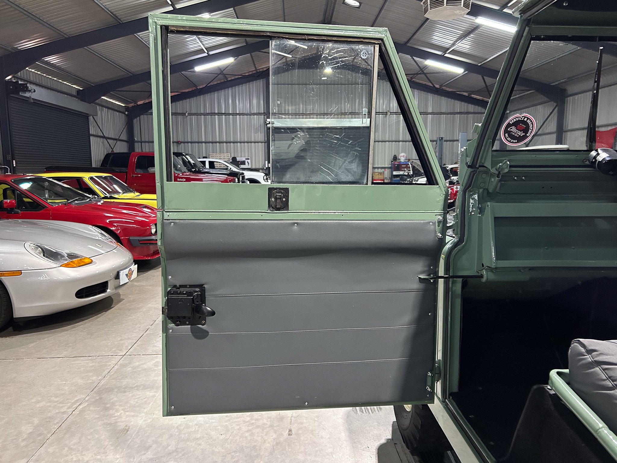 Indoor showroom or garage with a line of colorful classic cars in the background; in the foreground, a large green vehicle door is open, exposing a cracked window and metal paneling.