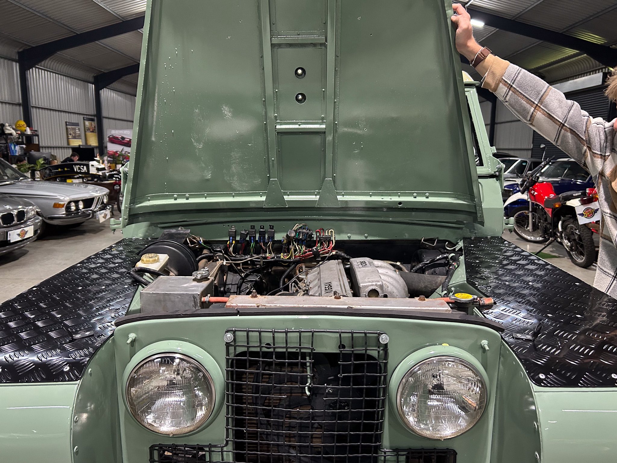 Open-hood view of olive-green vintage jeep in a workshop, showcasing exposed engine wiring and radiator grille.