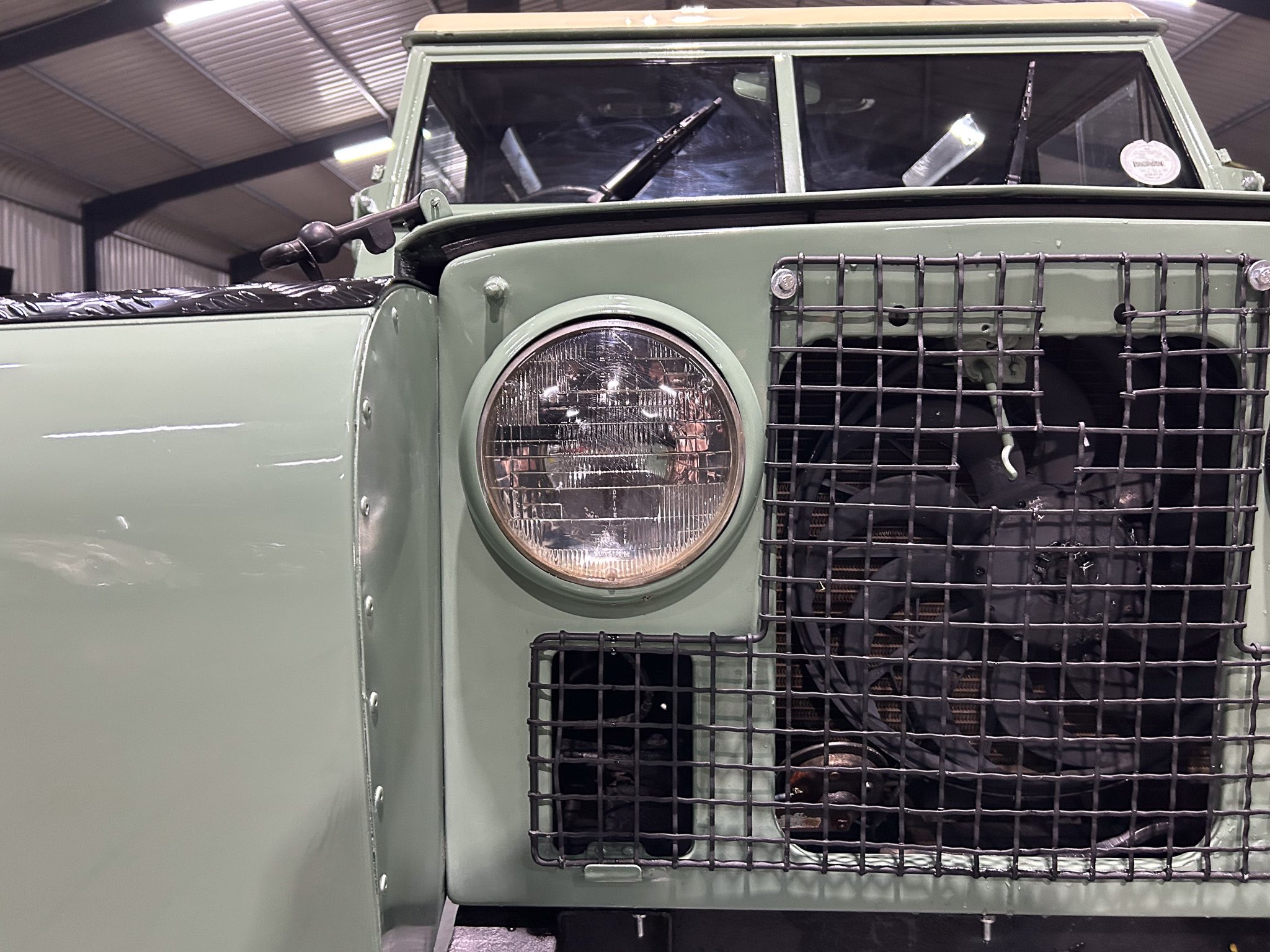 Close-up of the olive-green vehicle front with a metal protective grille over the grille and a round headlight visible on the left side inside a workshop.