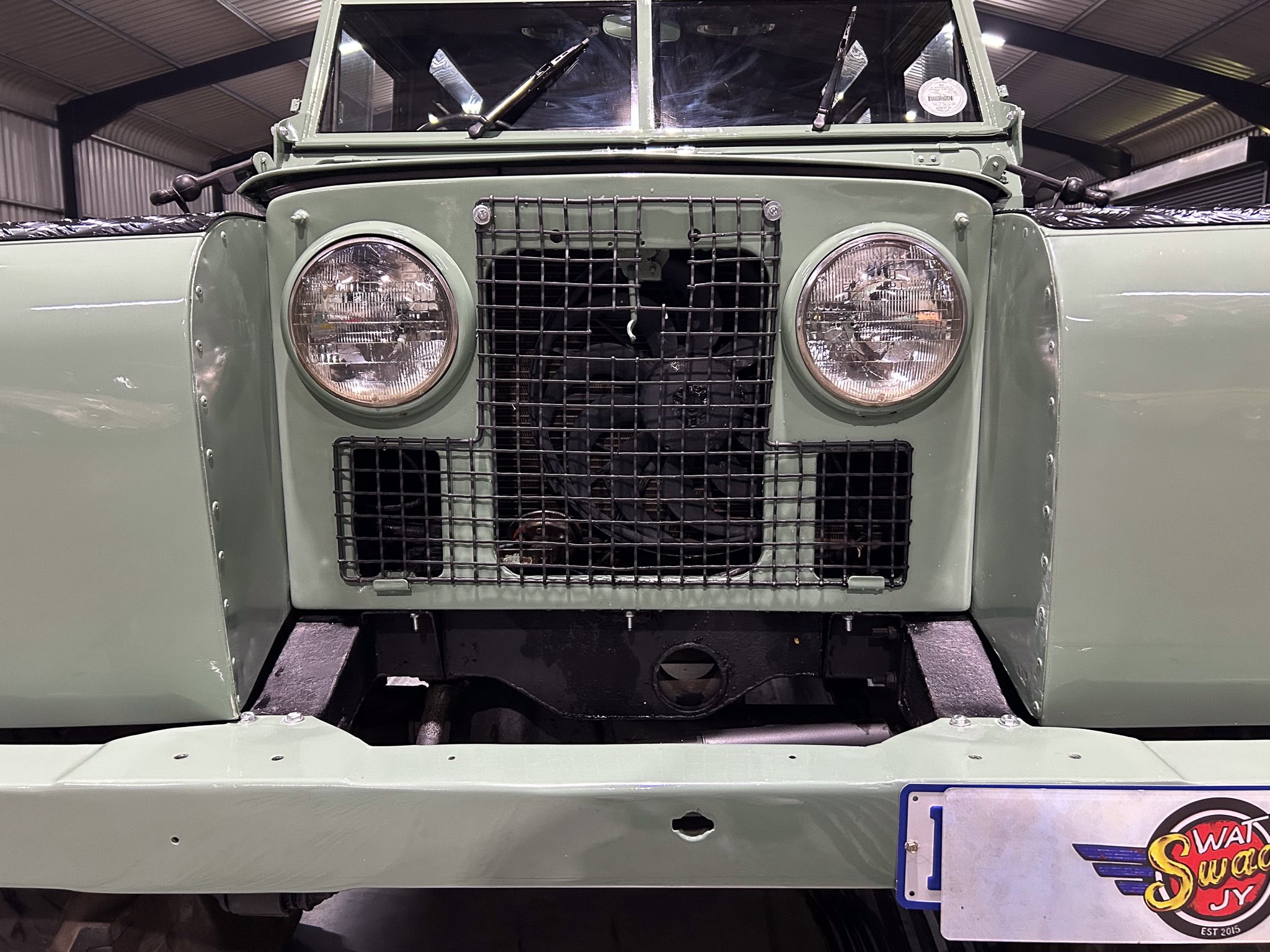 Front close-up of a vintage green military-style truck showing round headlights, protective grille, and open engine area in a warehouse setting.