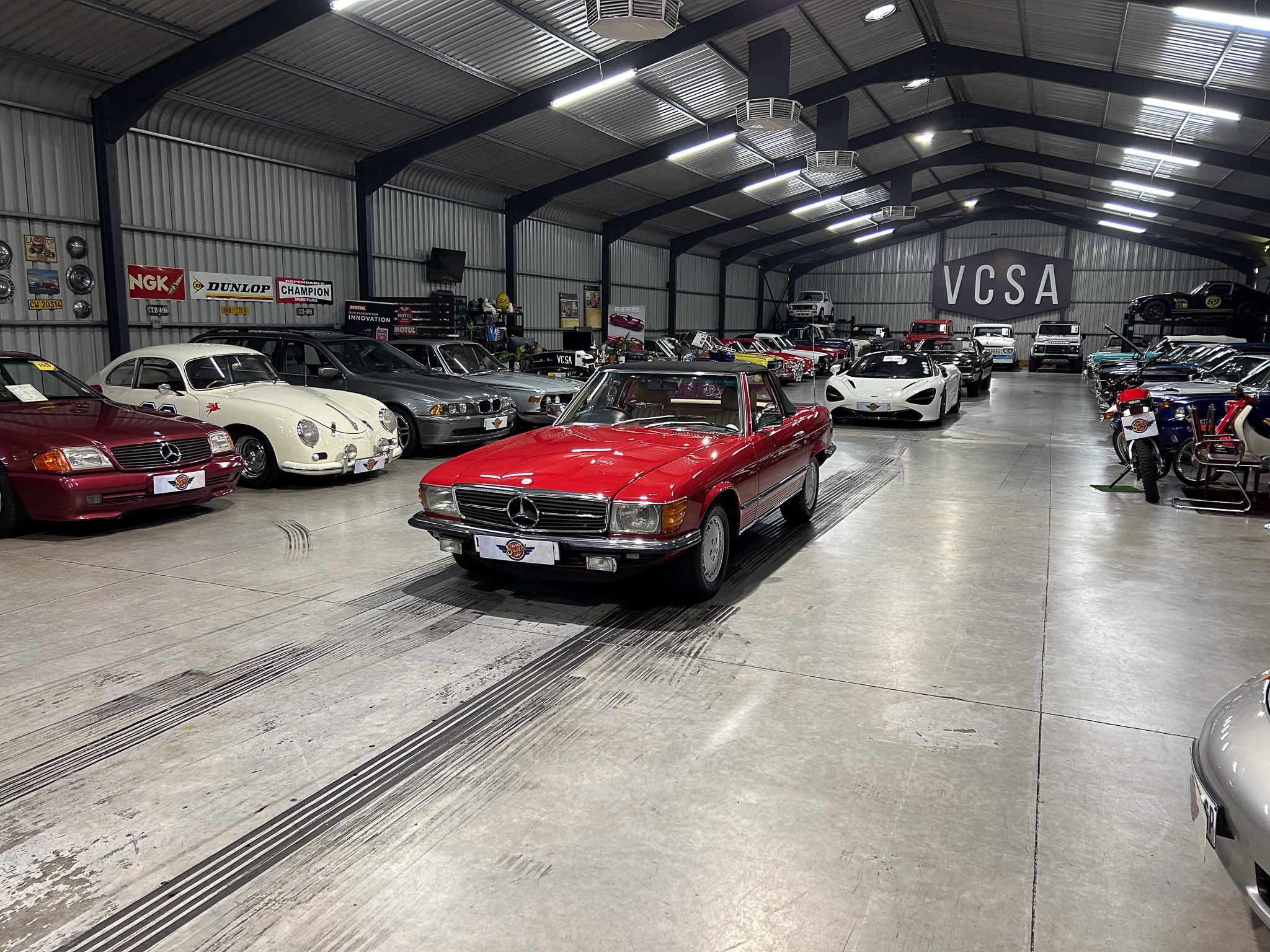 Interior of a large car showroom filled with vintage cars lined up along concrete floors under a metal warehouse roof with a VCSA sign in the background.