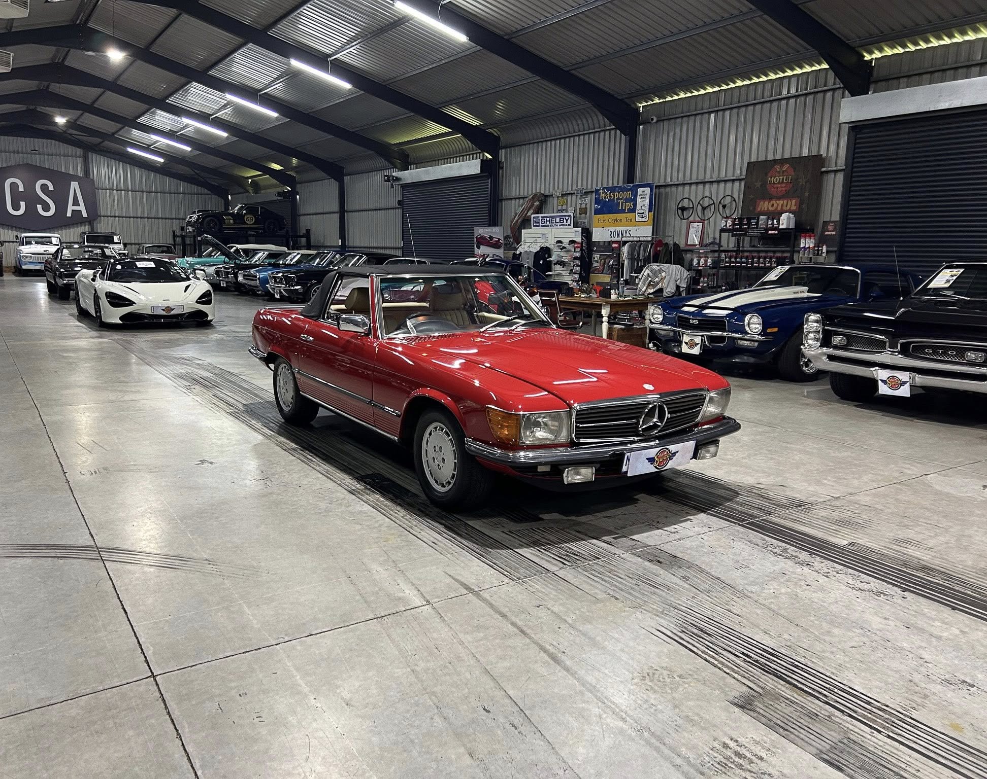 Indoor car showroom with a red vintage convertible in the foreground and rows of classic cars inside a metal warehouse.