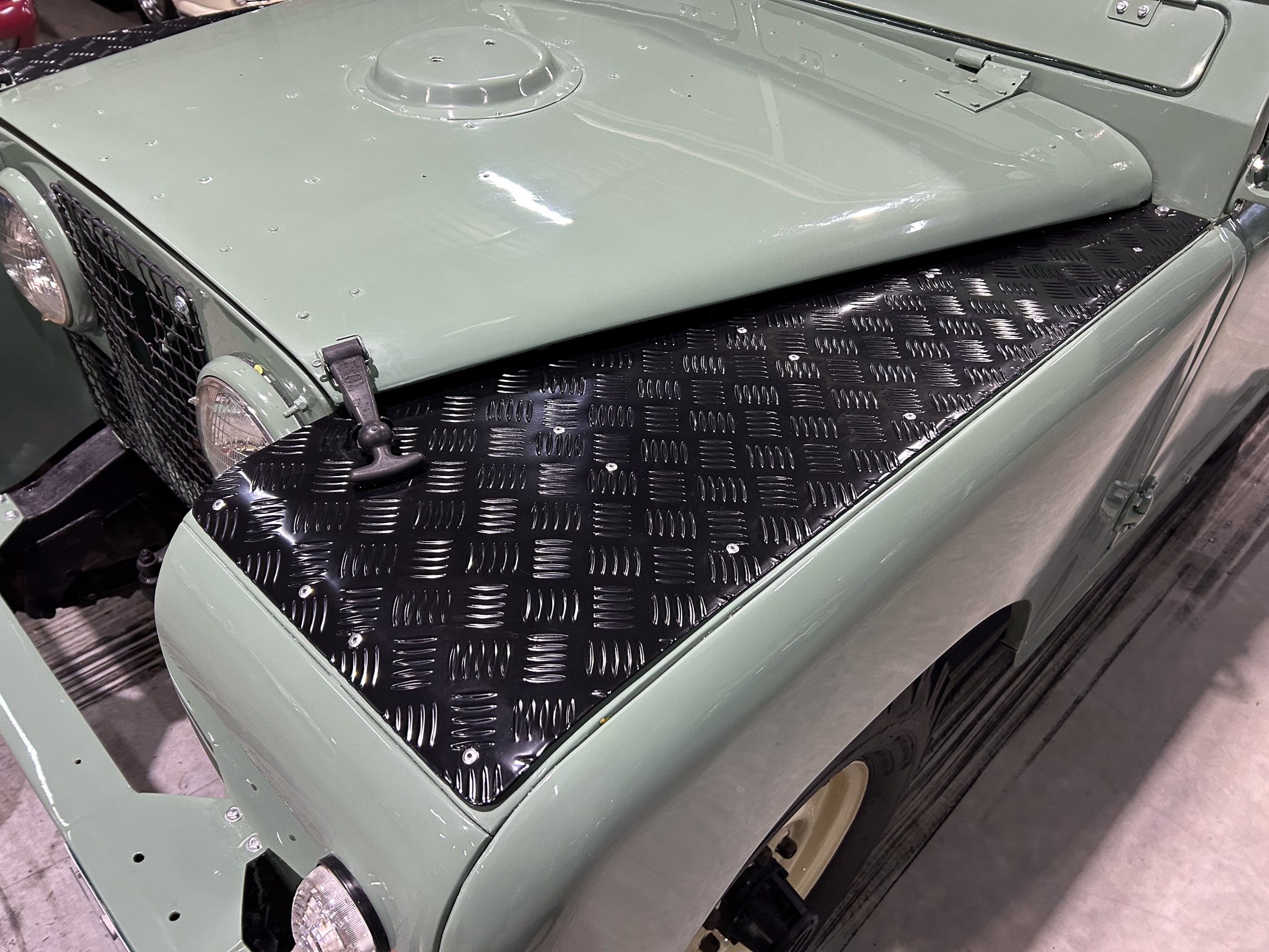 Front-quarter view of an olive-green vintage military jeep with a diamond-plate metal hood area.
