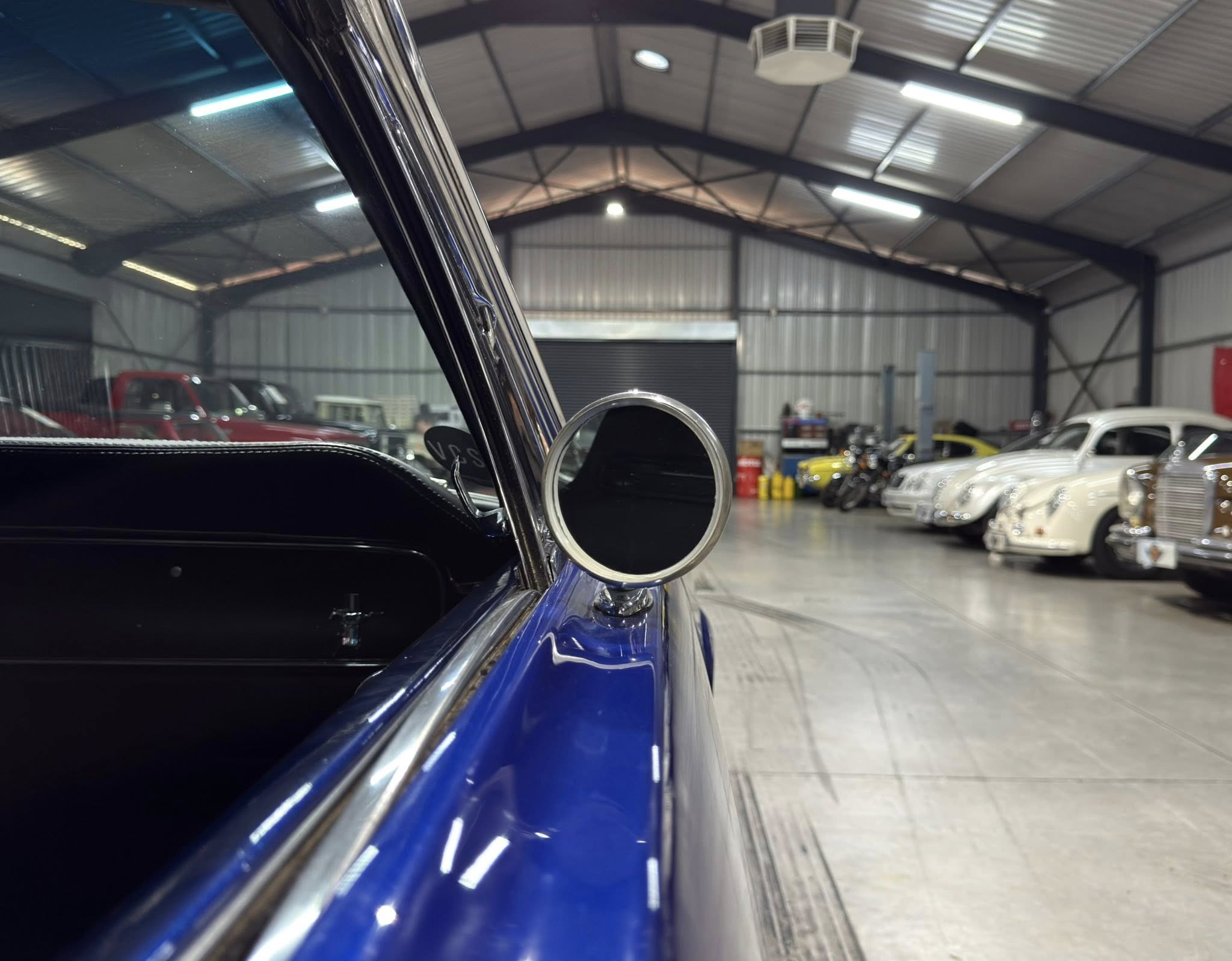 Close-up of a blue vintage car's side with chrome trim, inside a car showroom with other classic cars in the background.