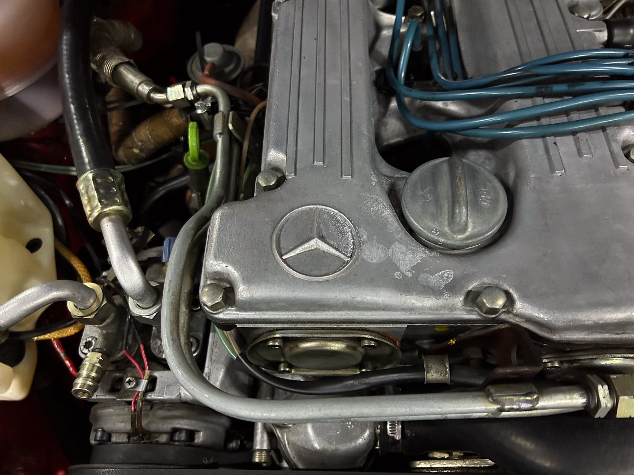 Close-up of a car engine bay showing a gray valve cover with the Mercedes-Benz star emblem, blue ignition wires, and various hoses and clamps surrounding it.