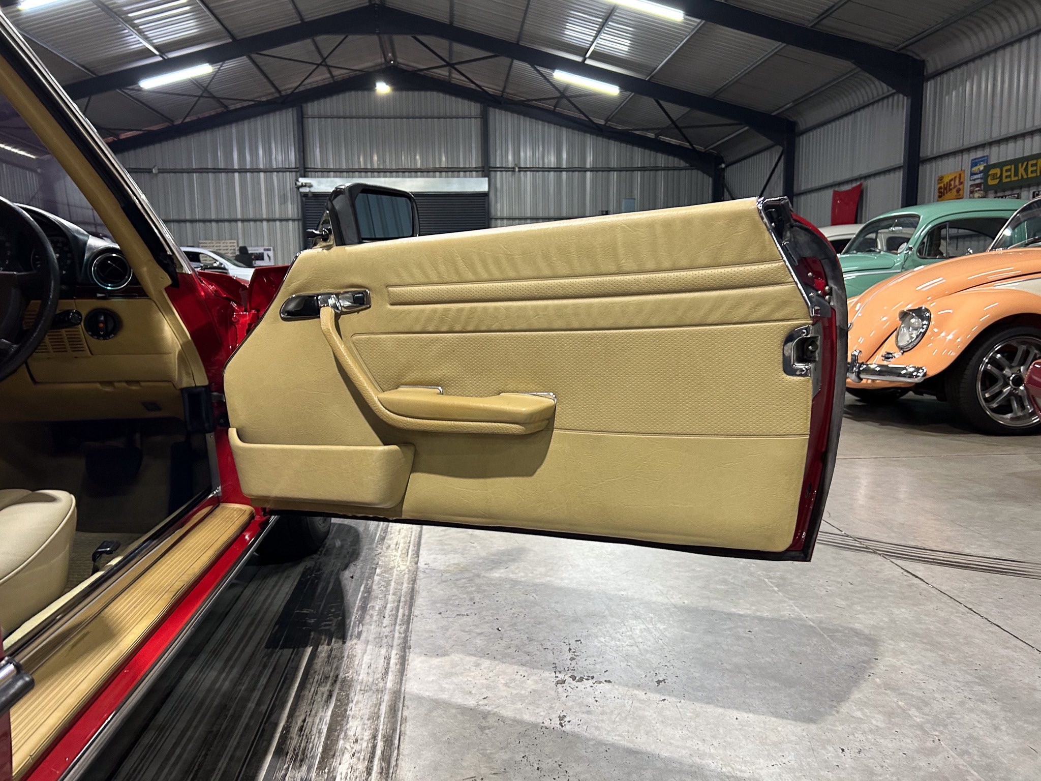 Open tan car door revealing beige interior in a classic car showroom with other vintage cars in the background, indoors.
