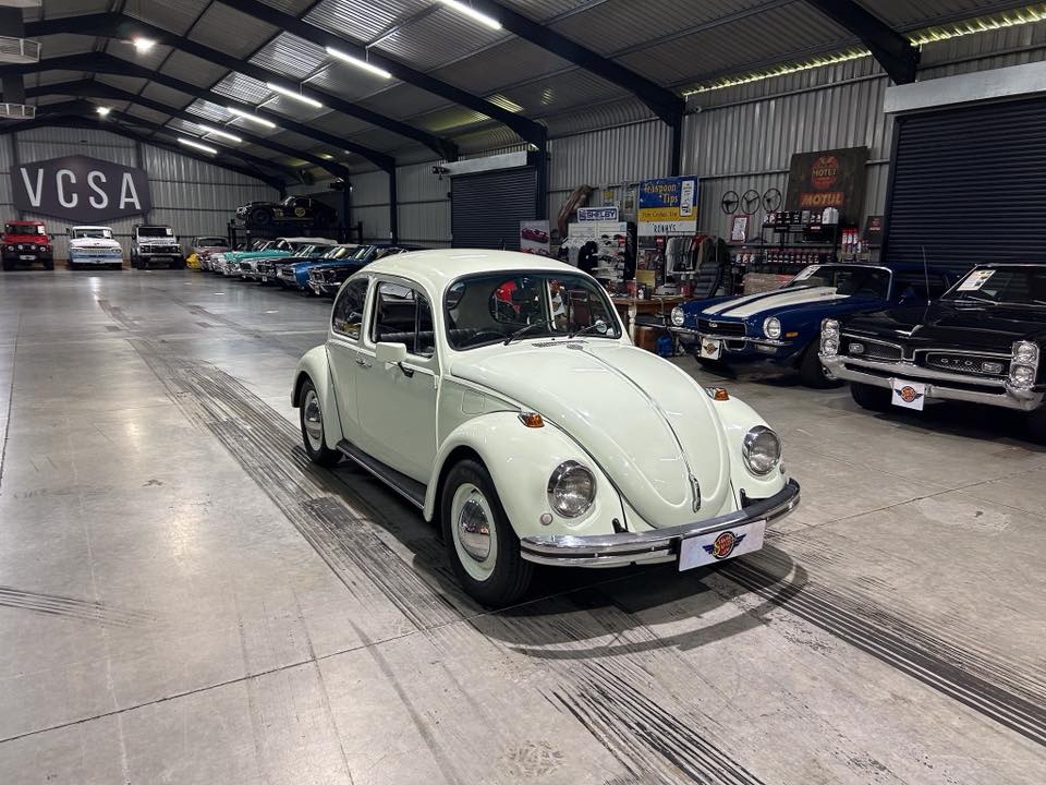 White vintage Volkswagen Beetle in a spacious showroom with other classic cars lined up along the walls and banners overhead, bright indoor lighting.