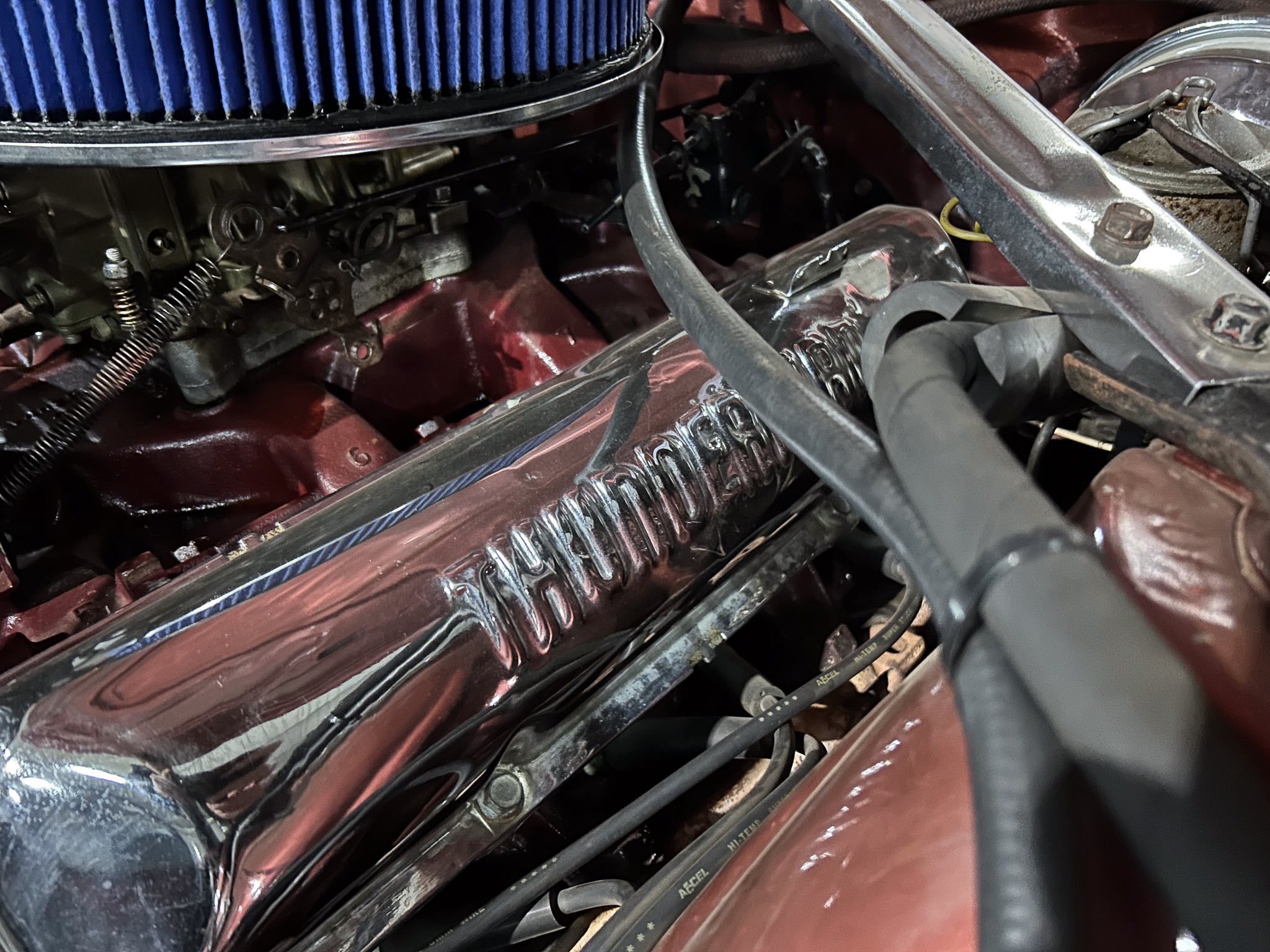 Close-up of a car engine bay showing a blue air filter, chrome valve cover, and red body with hoses and brackets around it.