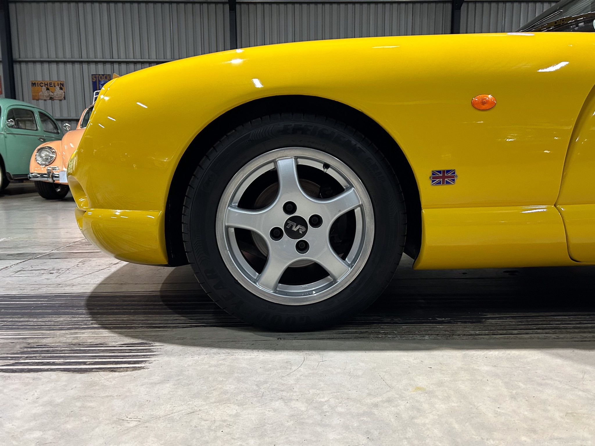 Close-up of a bright yellow sports car front wheel with alloy rim inside a museum showroom, other vintage cars in the background