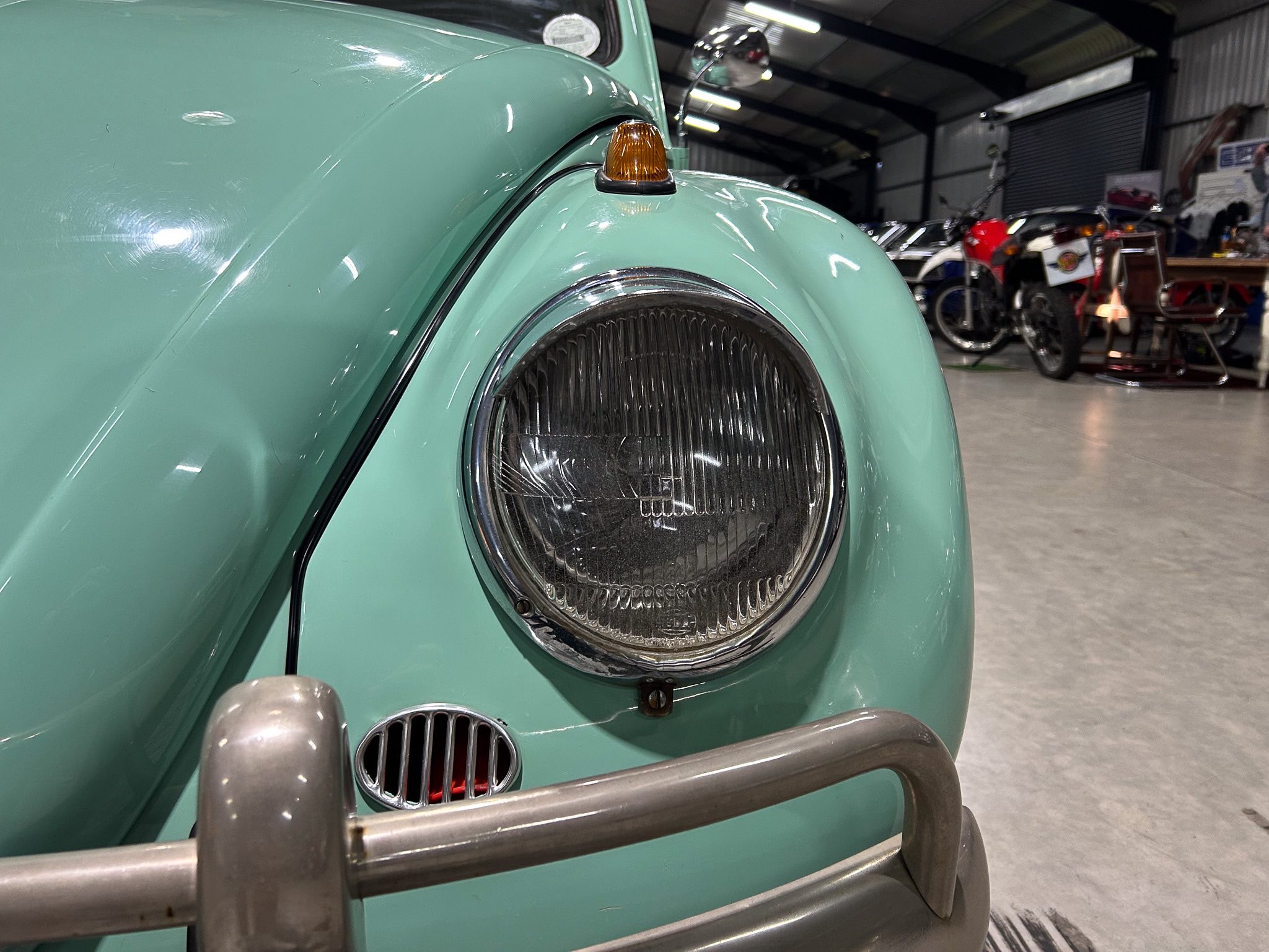 Close-up of a mint-green vintage car's headlight and orange indicator, chrome bumper visible in a showroom with motorcycles in the background.