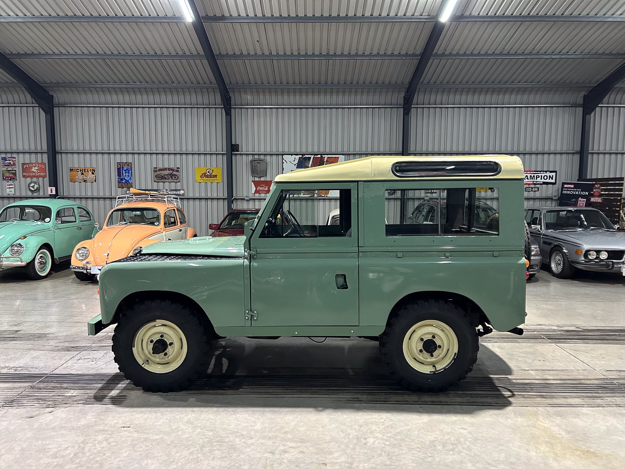 Green vintage utility vehicle in a museum-like showroom, with pastel cars along the wall and retro posters on a metal wall backdrop.