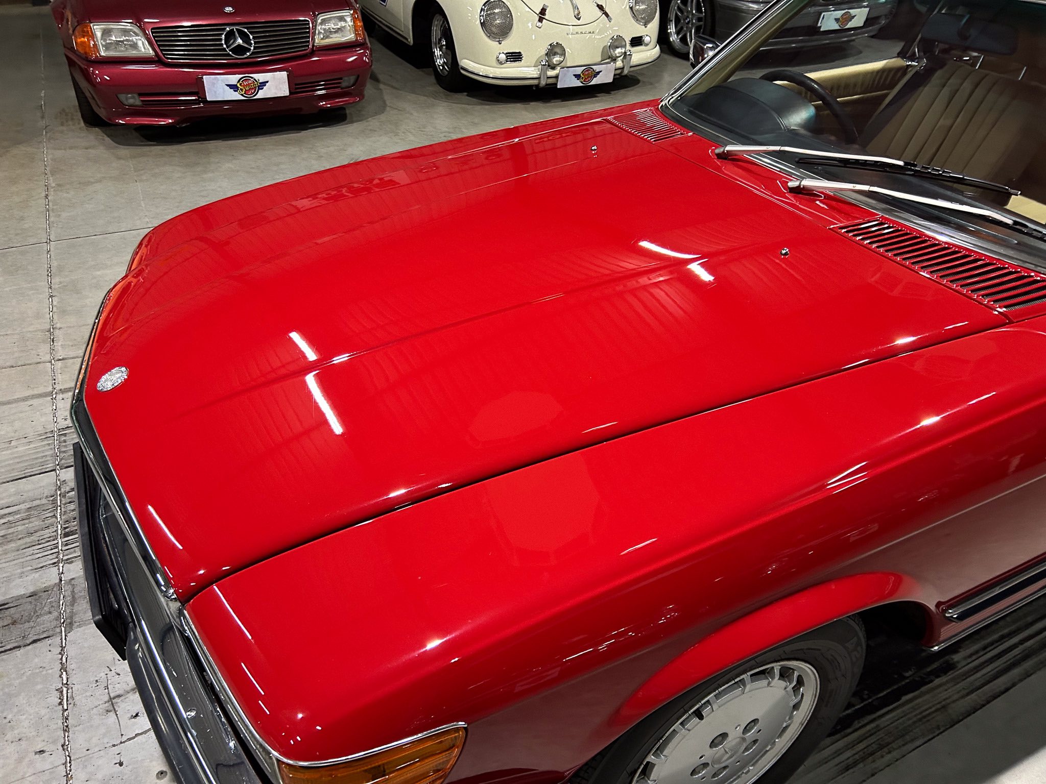 Close-up of a red vintage car hood and windshield wipers in a showroom with other classic cars in the background.