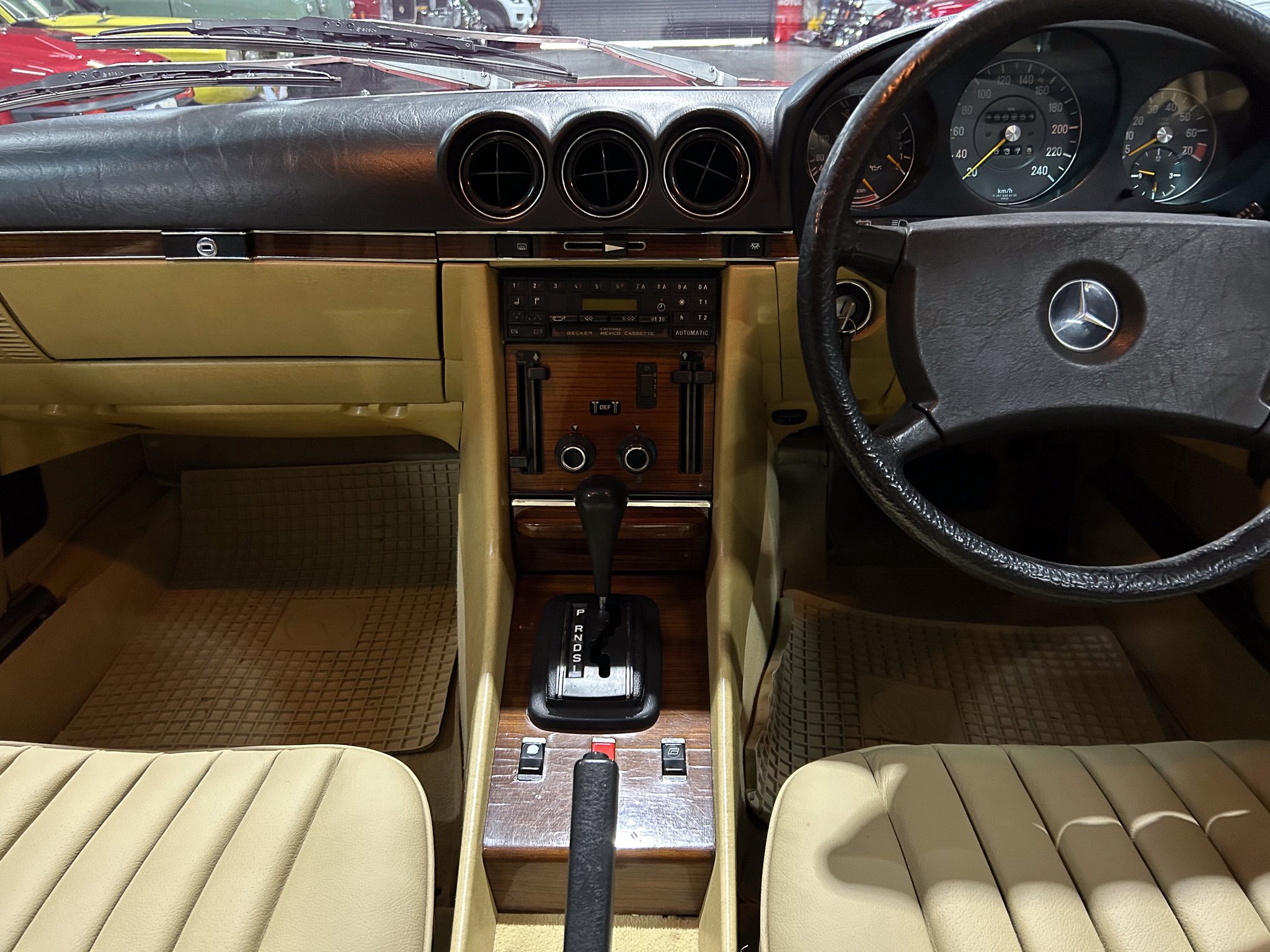 Interior of a vintage car showing tan seats, wood center console, three round air vents, and a black steering wheel with a Mercedes emblem.