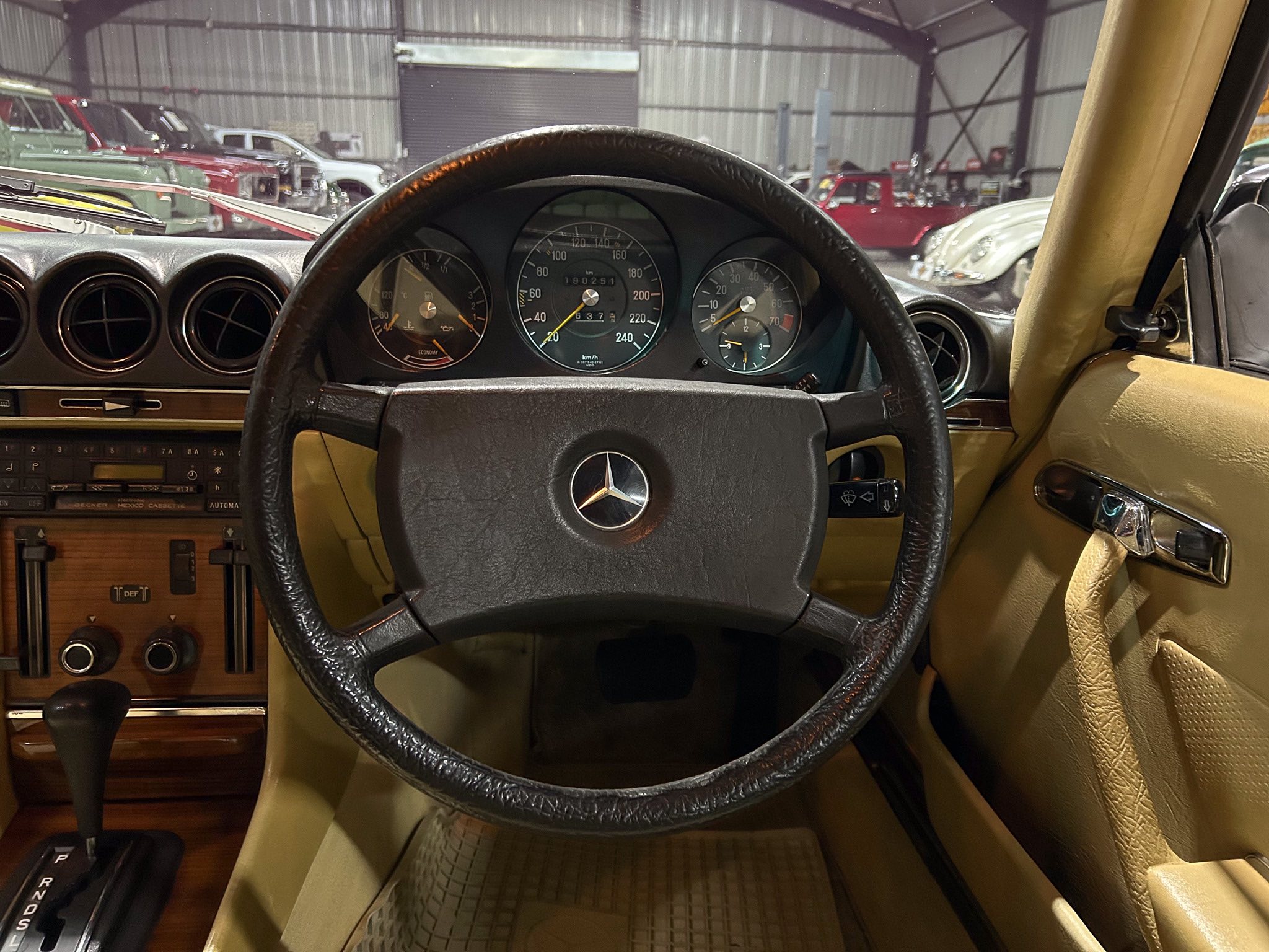 Driver's-eye view of a vintage car interior featuring a black leather steering wheel, analog gauges, and a tan dashboard with other classic cars visible outside the windshield in a showroom setting.