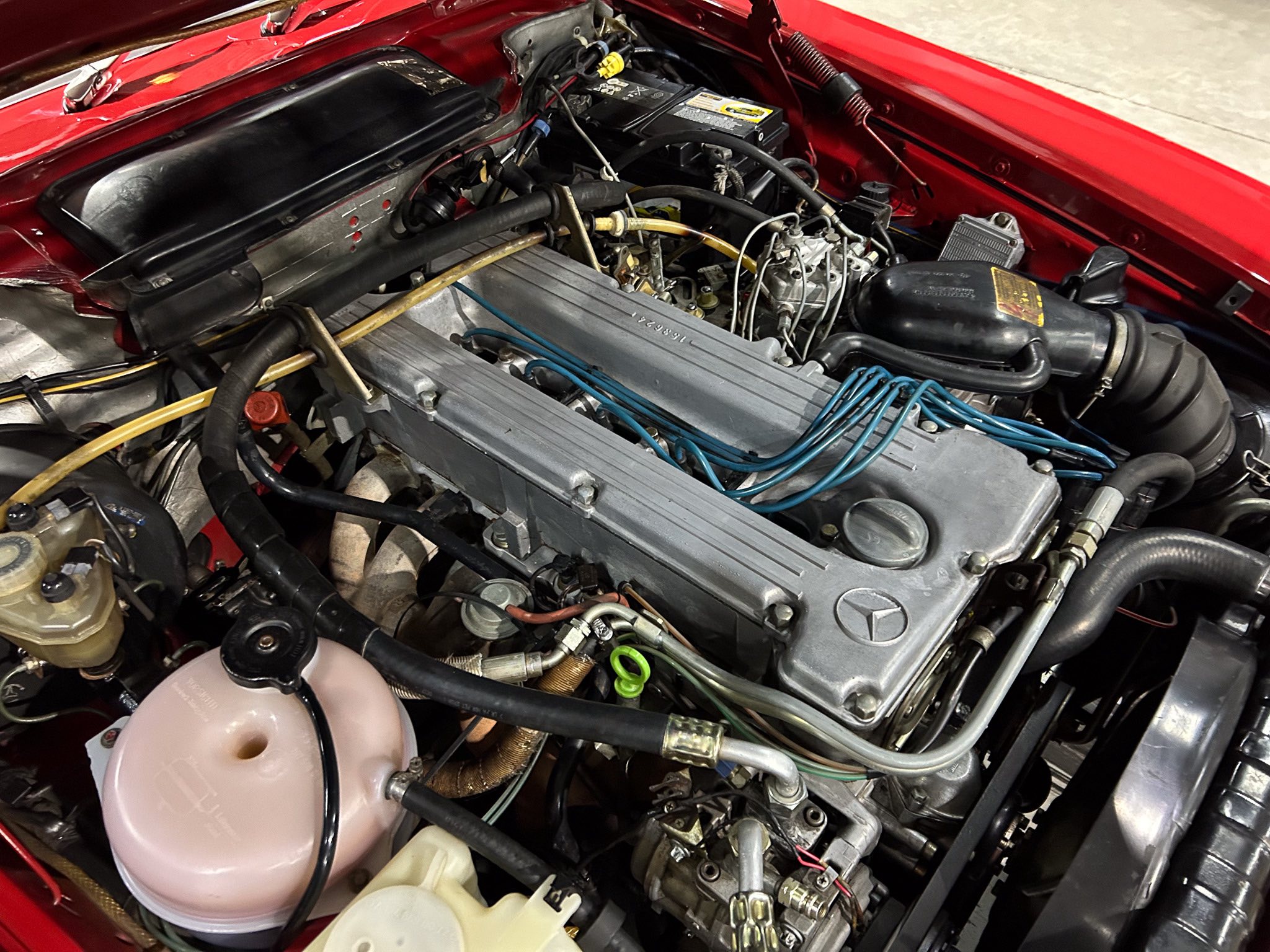 Close-up of a car engine bay with the battery, coolant reservoir, and numerous hoses and metal components visible in a red-painted engine bay
