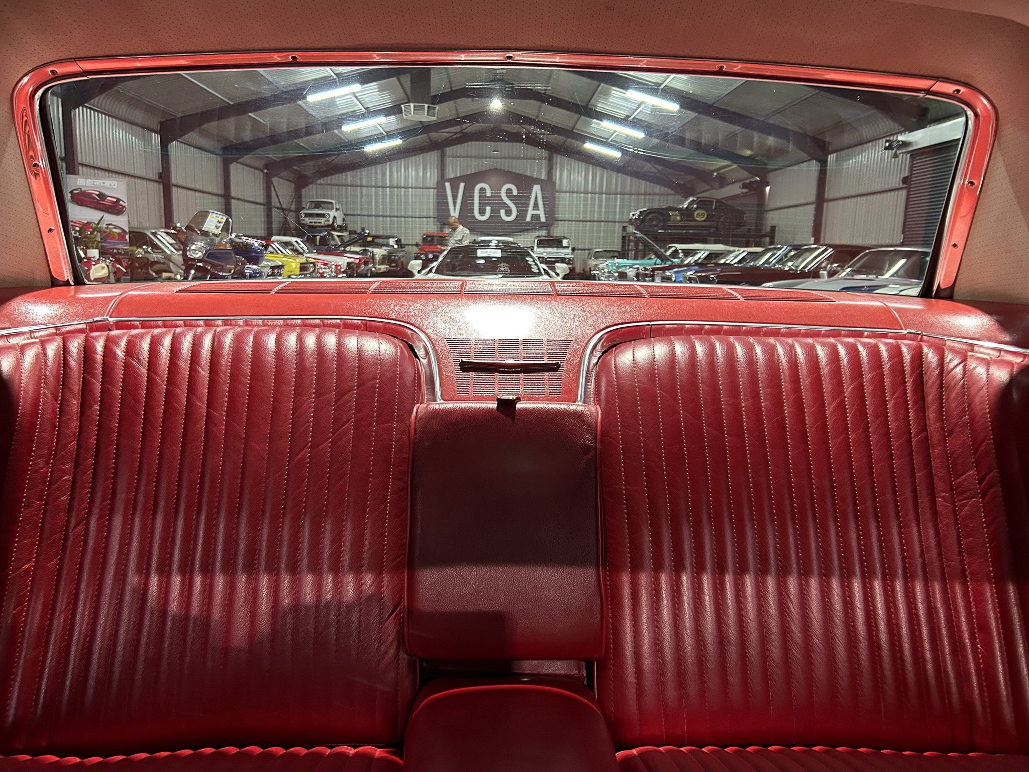 Red leather car interior viewed from the back seat, looking out at a showroom filled with classic cars and a VCSA sign.