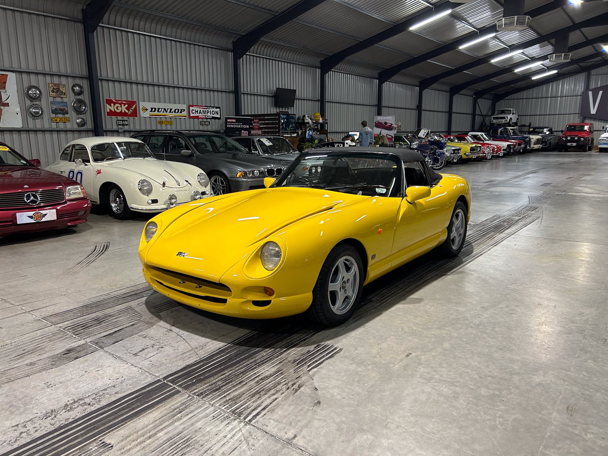 Inside a large metal warehouse filled with classic cars in a showroom lineup, the bright yellow convertible stands in the foreground.