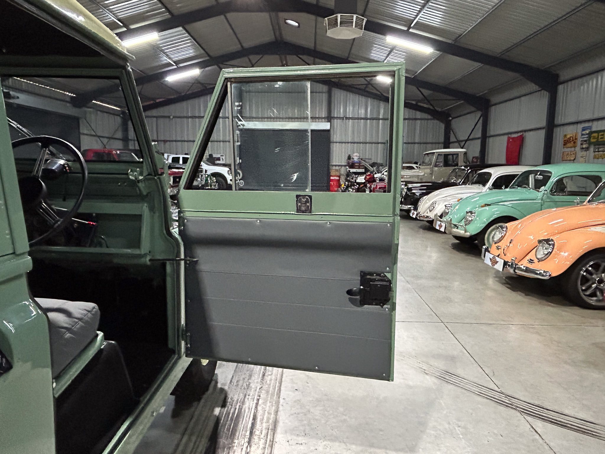 Open driver-side door of a green vintage vehicle in a museum or showroom, with classic cars lined up along the right side and a metal-roofed interior around.