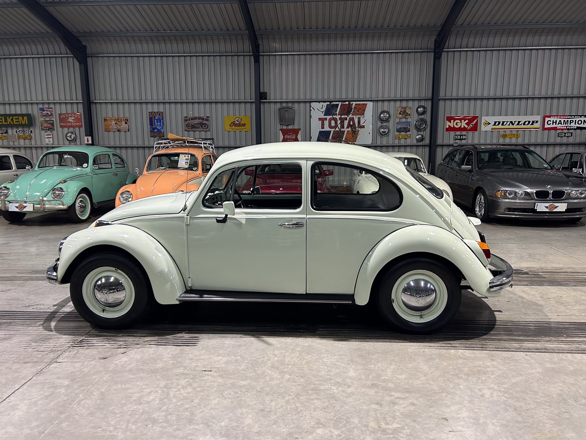 Side view of a white vintage Volkswagen Beetle in a showroom, with pastel-colored classic cars and retro signs on a corrugated metal wall behind them.