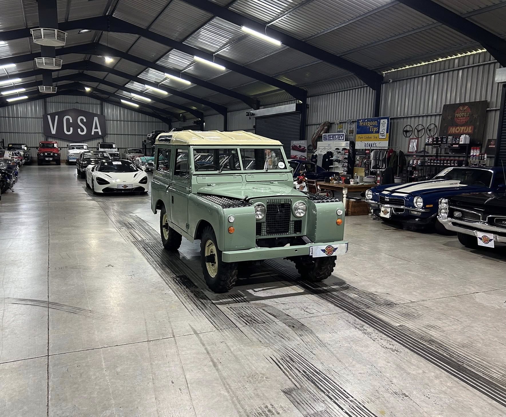 Indoor showroom filled with classic and modern cars, front-and-center pale green vintage off-road vehicle amid other exotics and memorabilia.
