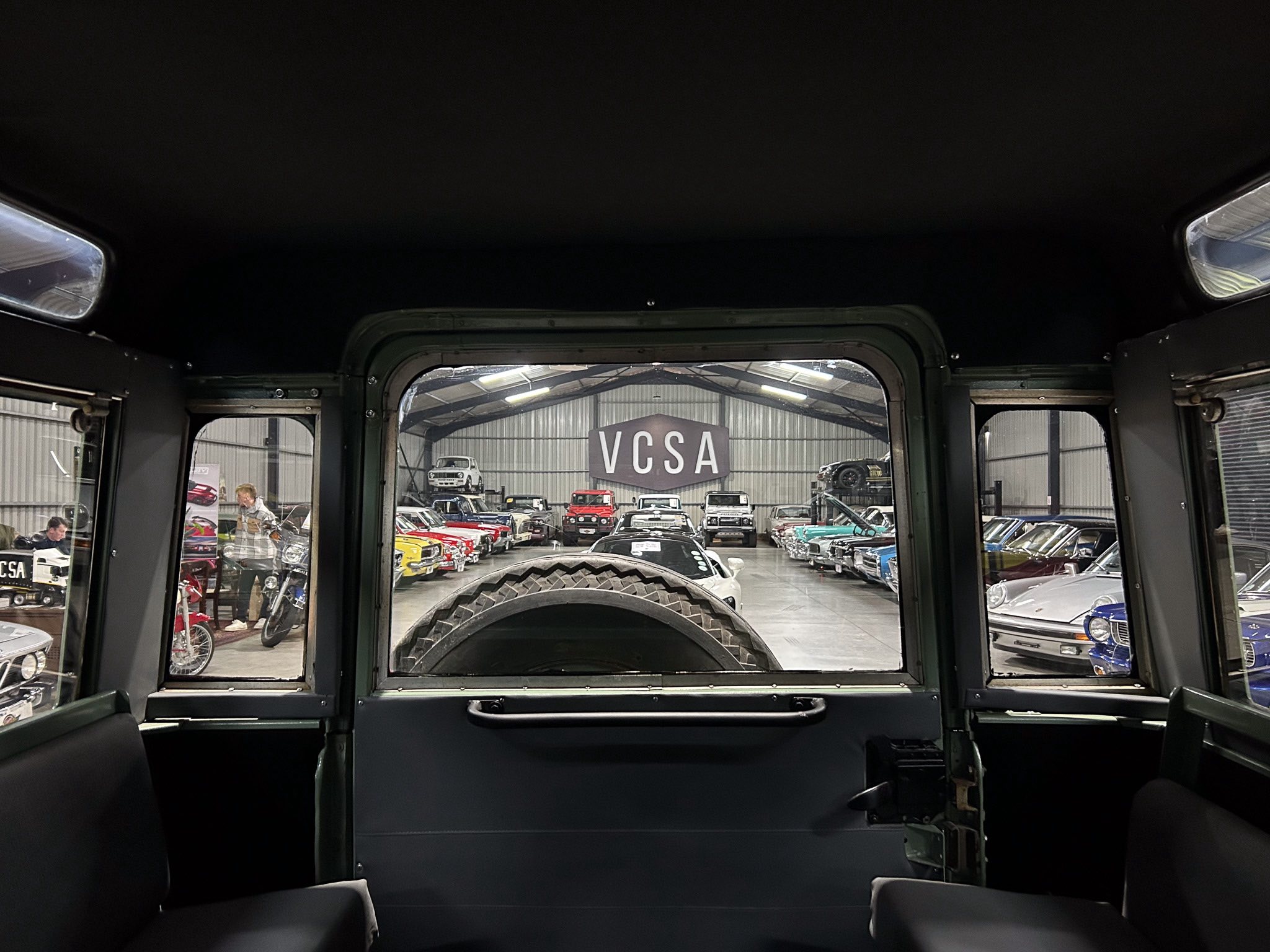 View from inside a vintage vehicle looking out at a showroom filled with classic cars and a large 'VCSA' sign in the background