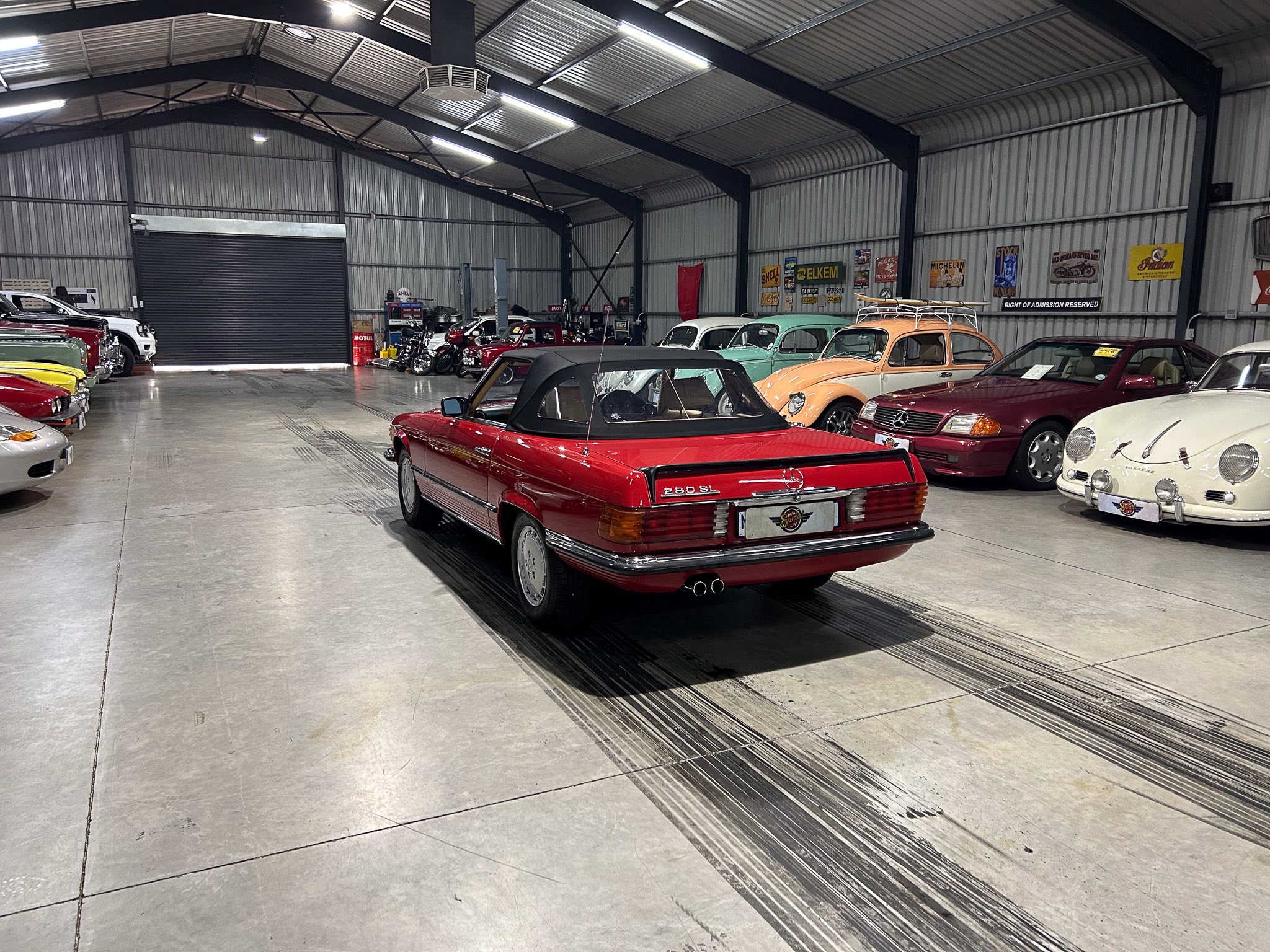 Indoor showroom filled with a row of colorful vintage cars, including a red convertible in the foreground and classic sedans along the sides in a metal warehouse setting.