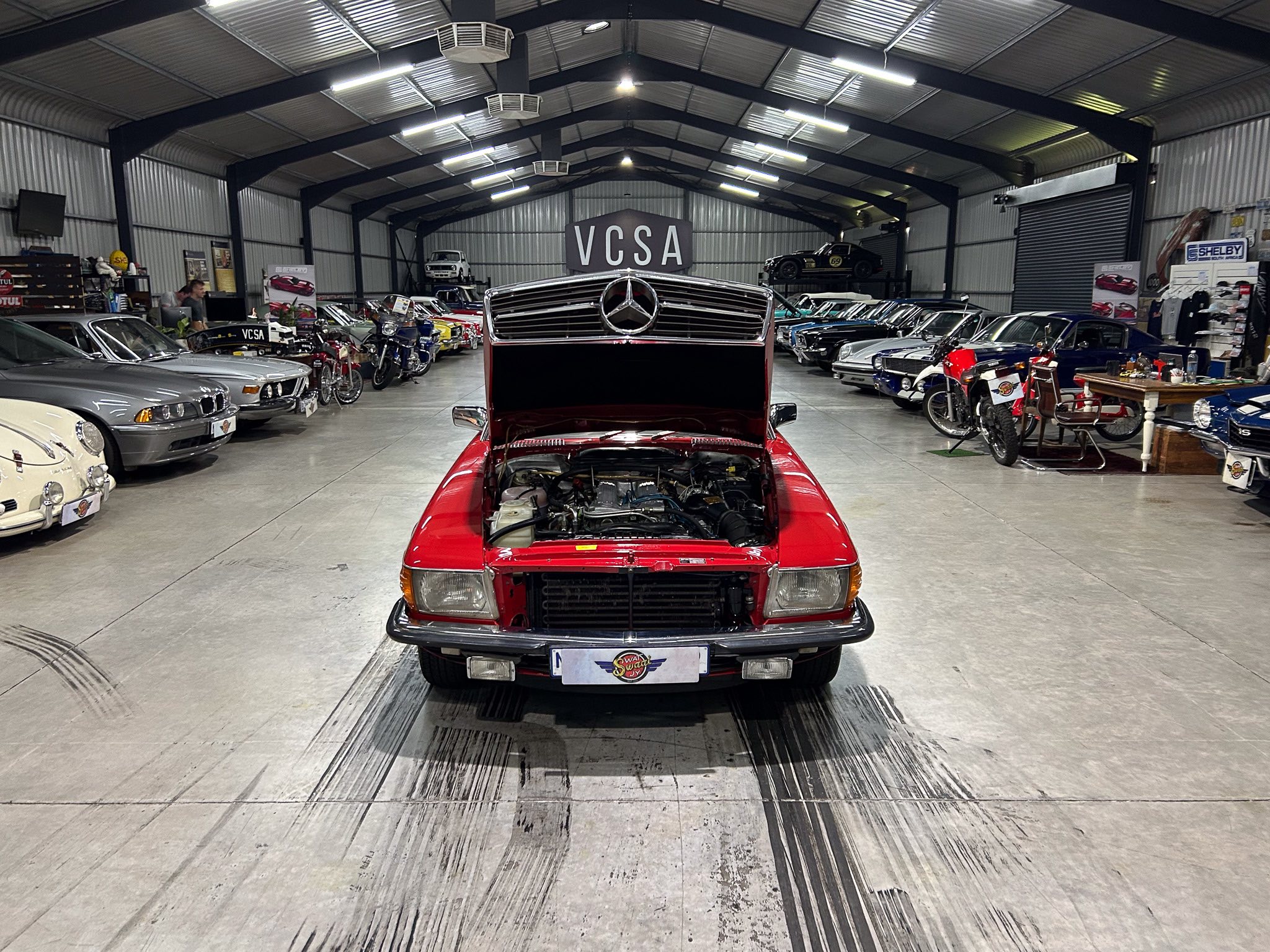 Mercedes-Benz show car in a large indoor showroom filled with classic cars and motorcycles, with a VC SA sign in the background.