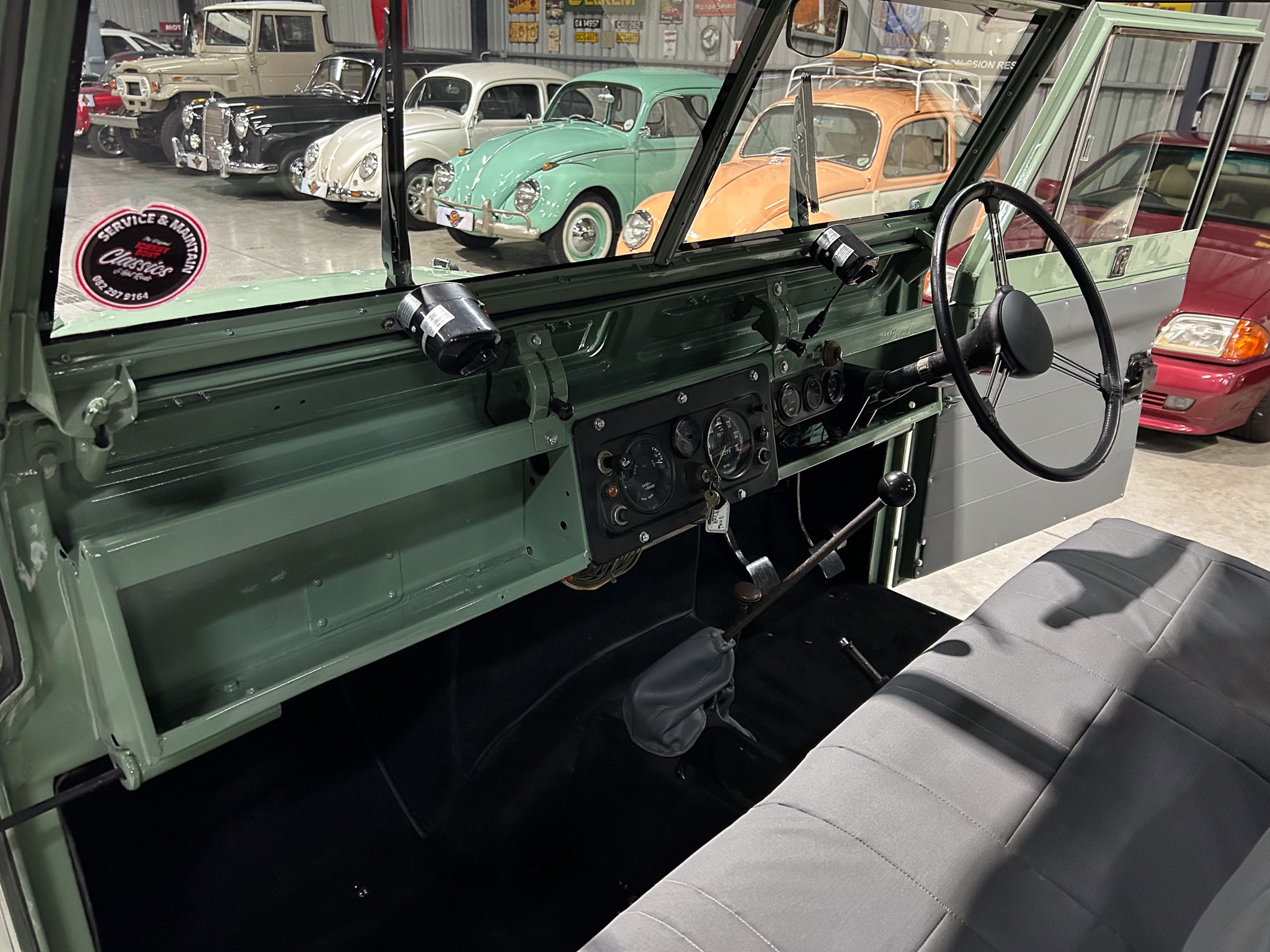 Interior of a vintage green vehicle showing steering wheel, gauges, and bench seat, with a classic car showroom in the background.