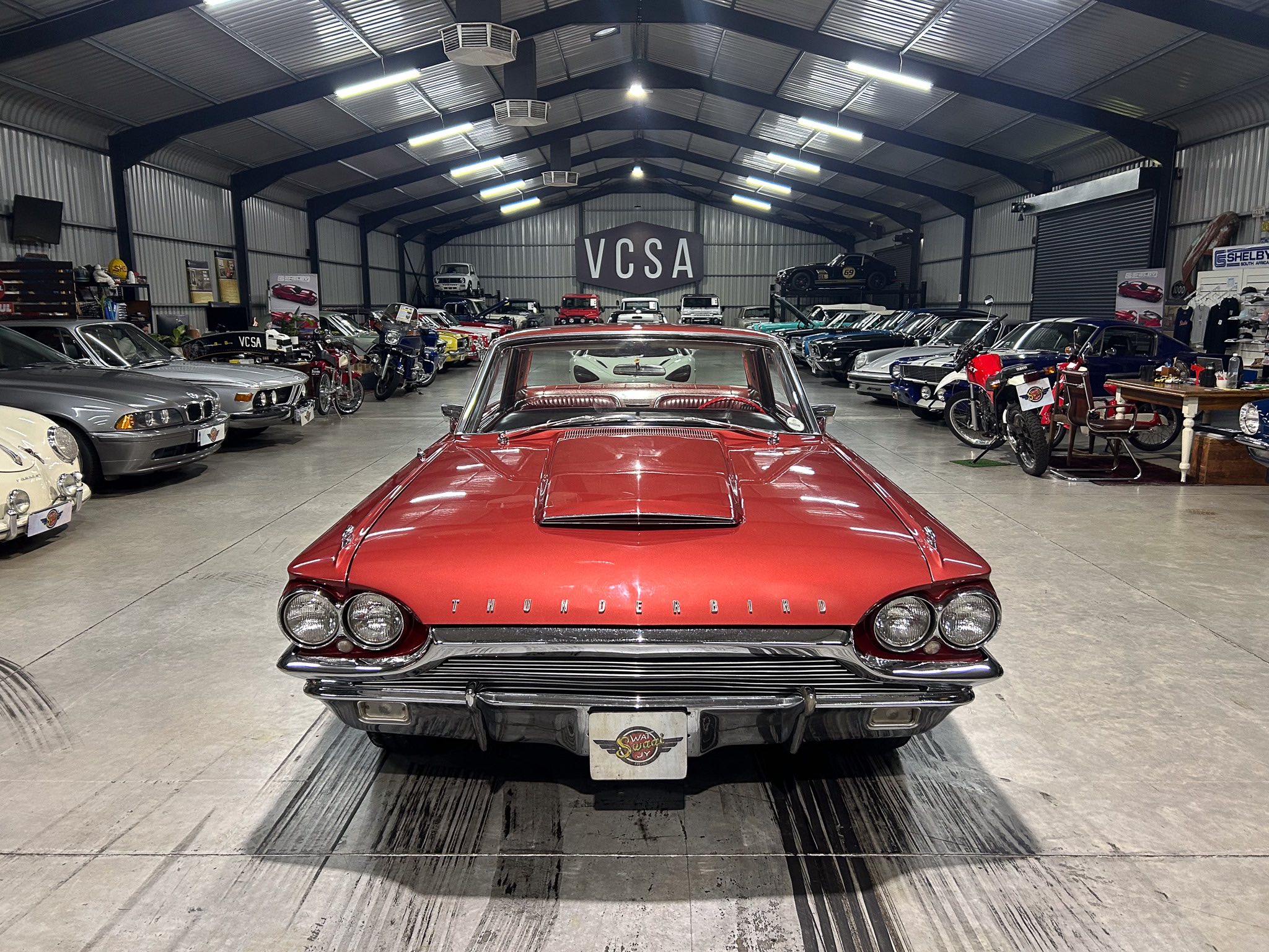 Red 1960s Ford Thunderbird parked center in a large well-lit indoor showroom filled with classic cars on both sides.