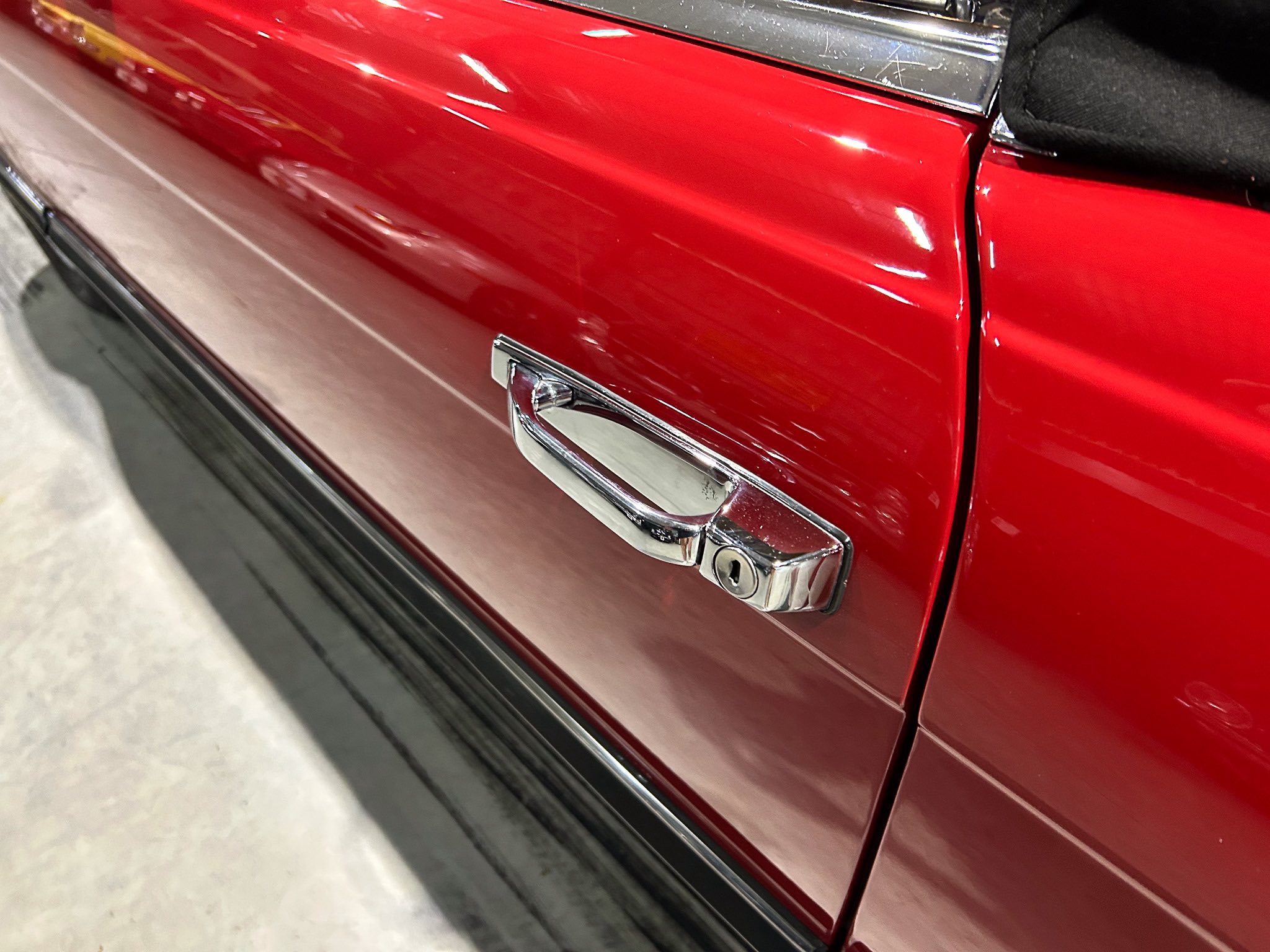 Close-up of a glossy red car door with a chrome door handle and black convertible top edge visible.