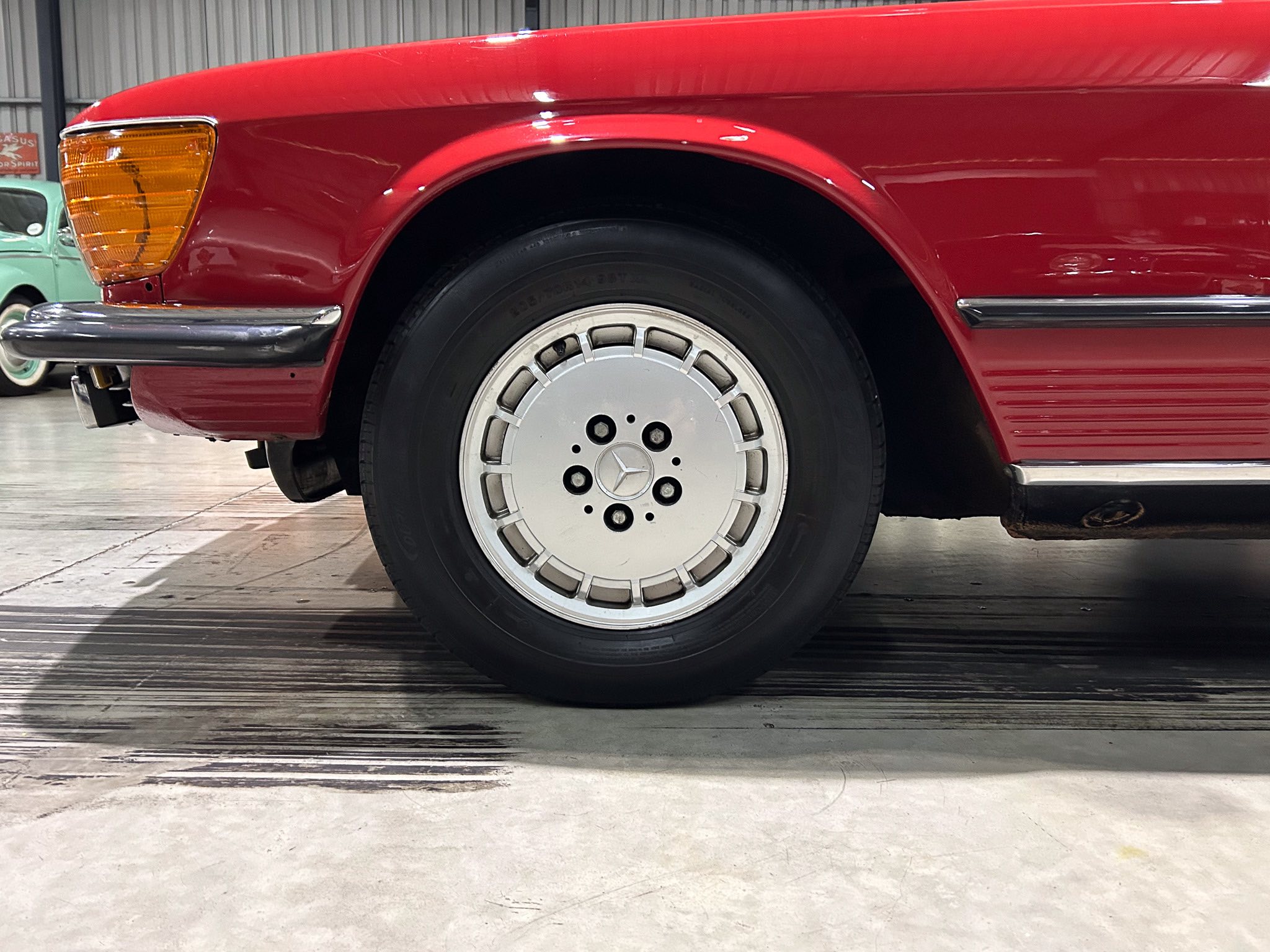 Close-up of a red vintage car's front left wheel and fender, with a chrome bumper and orange headlight visible in the background