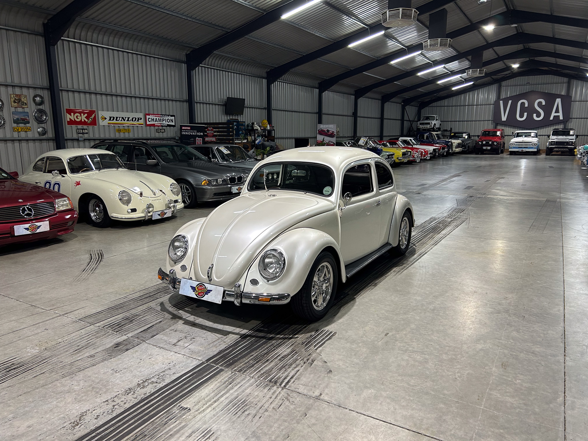 Indoor showroom with a row of vintage cars, foregrounded by a white classic VW Beetle and a long line of colorful classics under a metal roof with a VCSA sign in the background