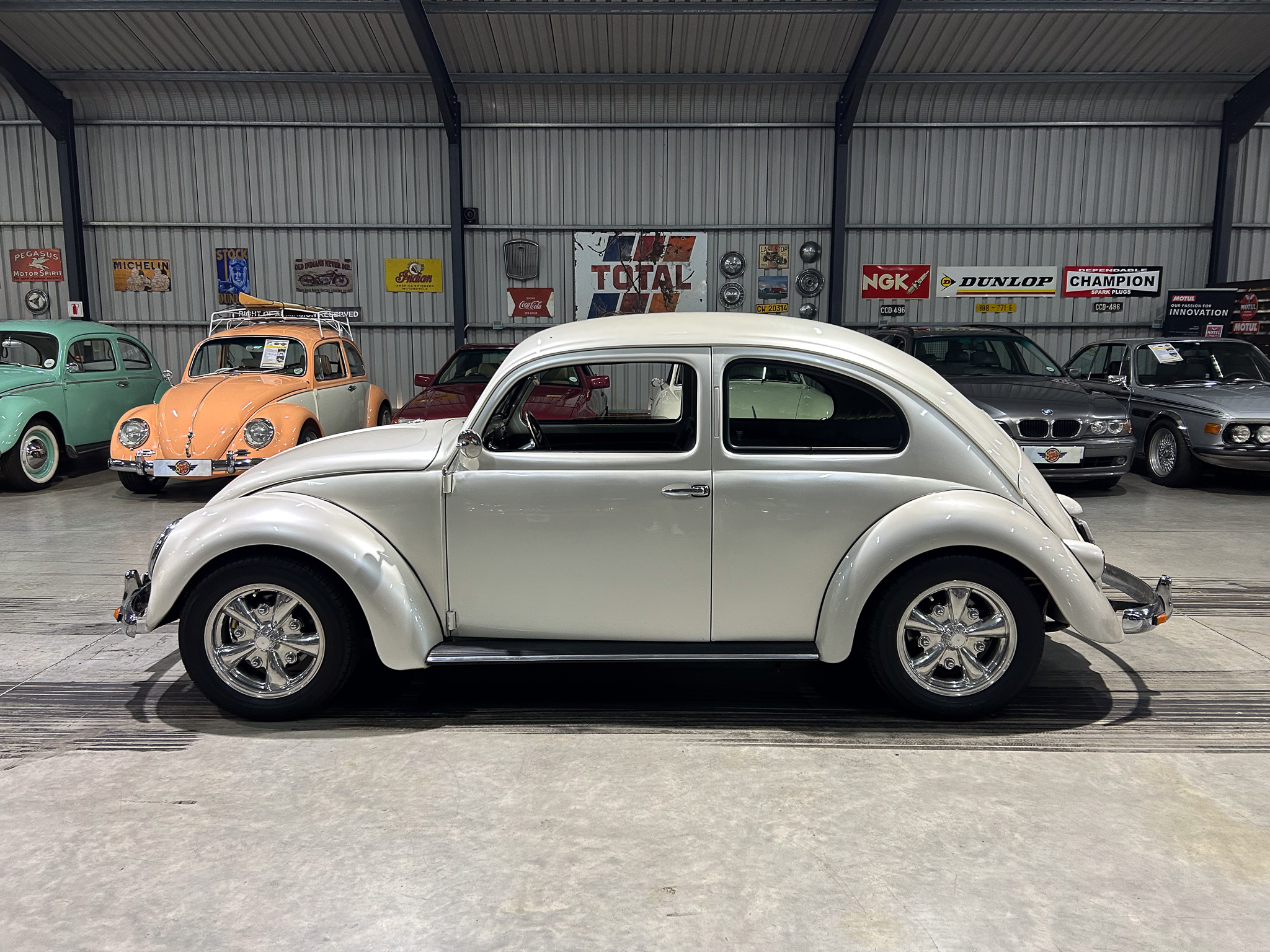White vintage car in a showroom with other classic cars and retro brand signs on a corrugated metal wall backdrop.